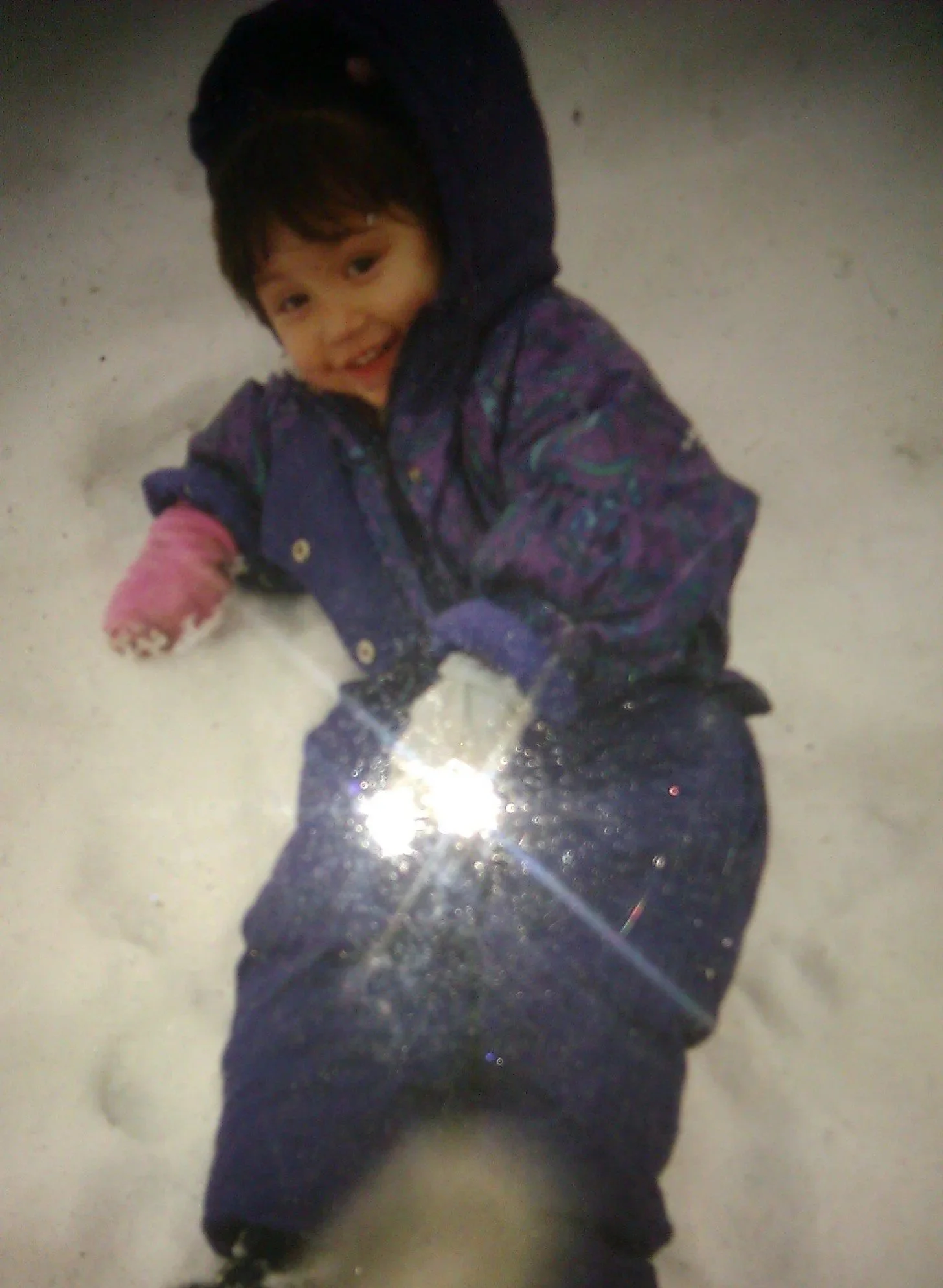 A young child with brown hair smiling and lying on the snow, wearing a purple and pink jacket and black snow pants.