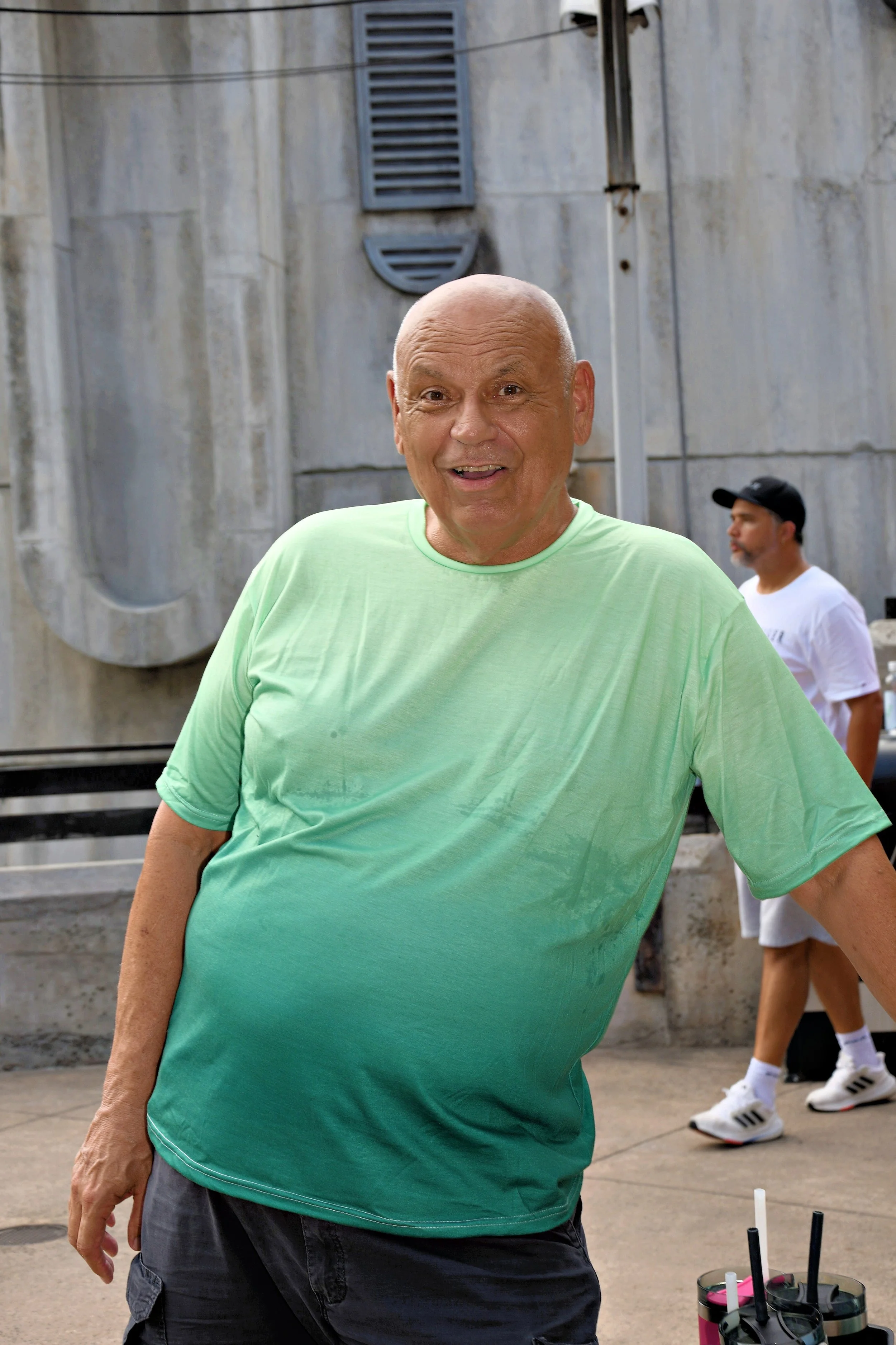 A cheerful bald man wearing a light green T-shirt and dark shorts stands outdoors with a concrete wall and a man in the background. The background includes a vent and a black pipe on the wall, and a drink container with straws is in the foreground.