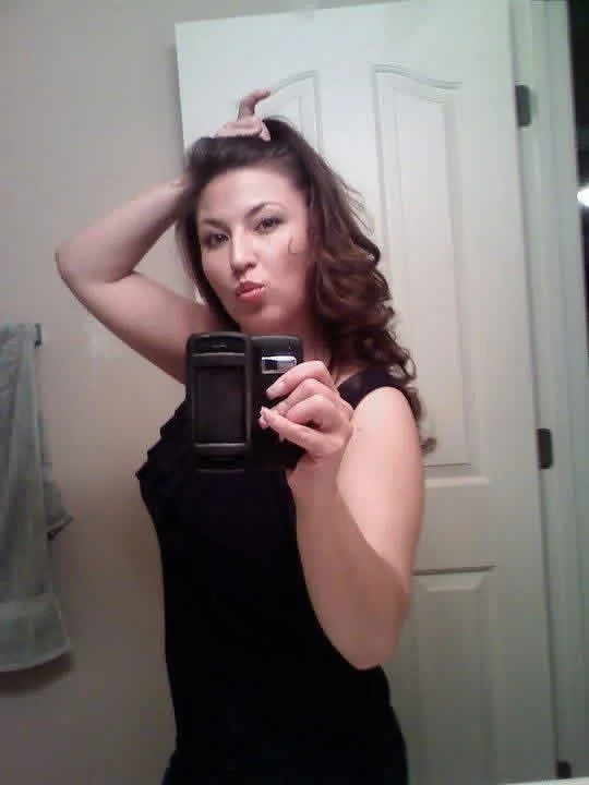 Woman taking a selfie in mirror, with hand on her head, wearing a black sleeveless top.