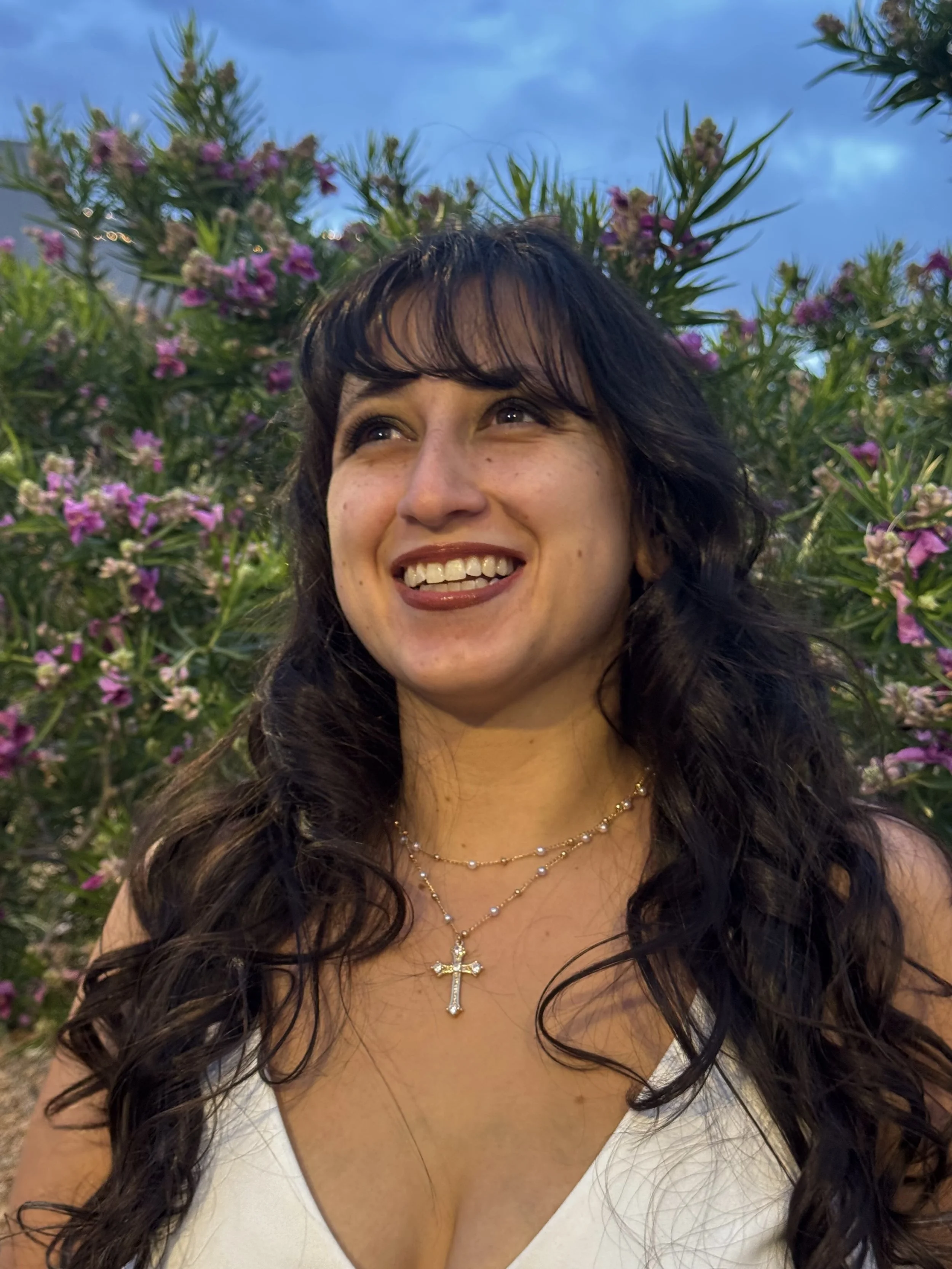 A woman with long dark curly hair is smiling and looking upward. She is wearing a white top with a low neckline, a gold necklace with a cross pendant, and has a background of pink and purple flowers with a blue sky.