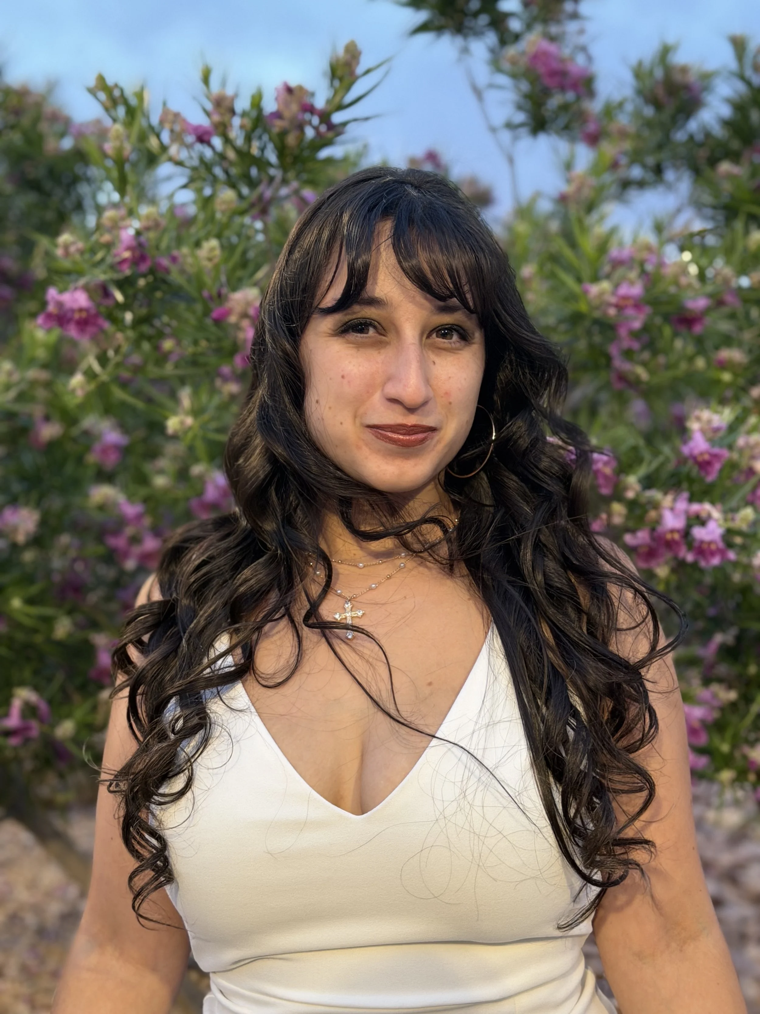 Close-up of a woman with long dark curly hair, wearing a white sleeveless top and a silver necklace with a cross pendant, standing outdoors with pink flowers and green foliage in the background.