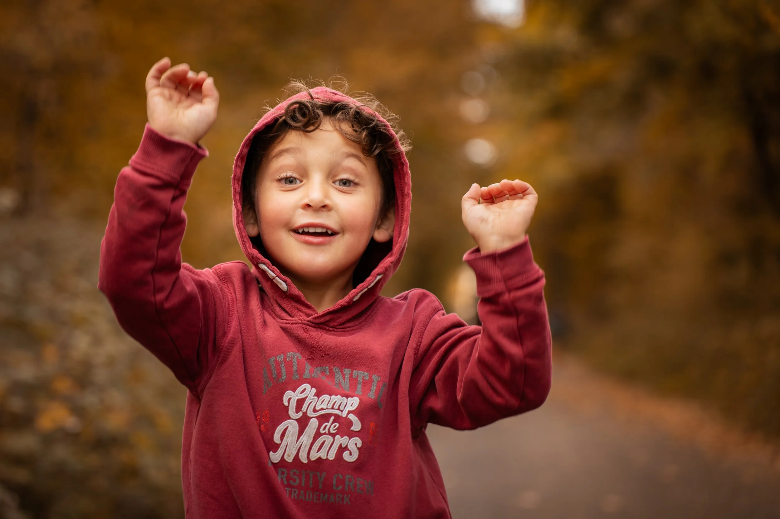 Un enfant souriant portant un sweat à capuche rouge, levant les bras, dans un environnement extérieur automnal avec des feuilles jaunes et marron.