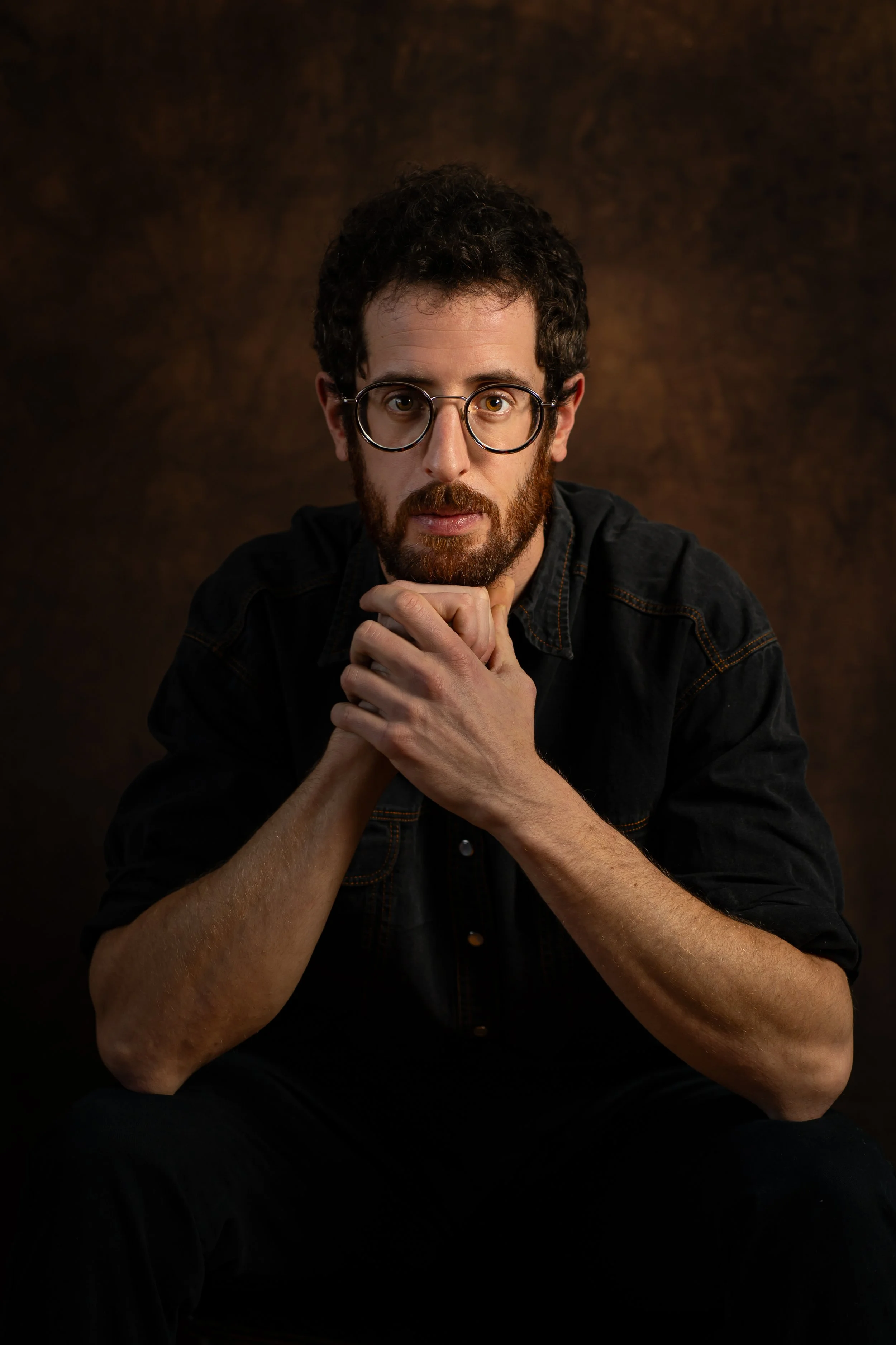 Un homme aux cheveux bouclés et à la barbe porte des lunettes rondes, regarde directement l'objectif, avec les mains sous le menton, contre un fond sombre.