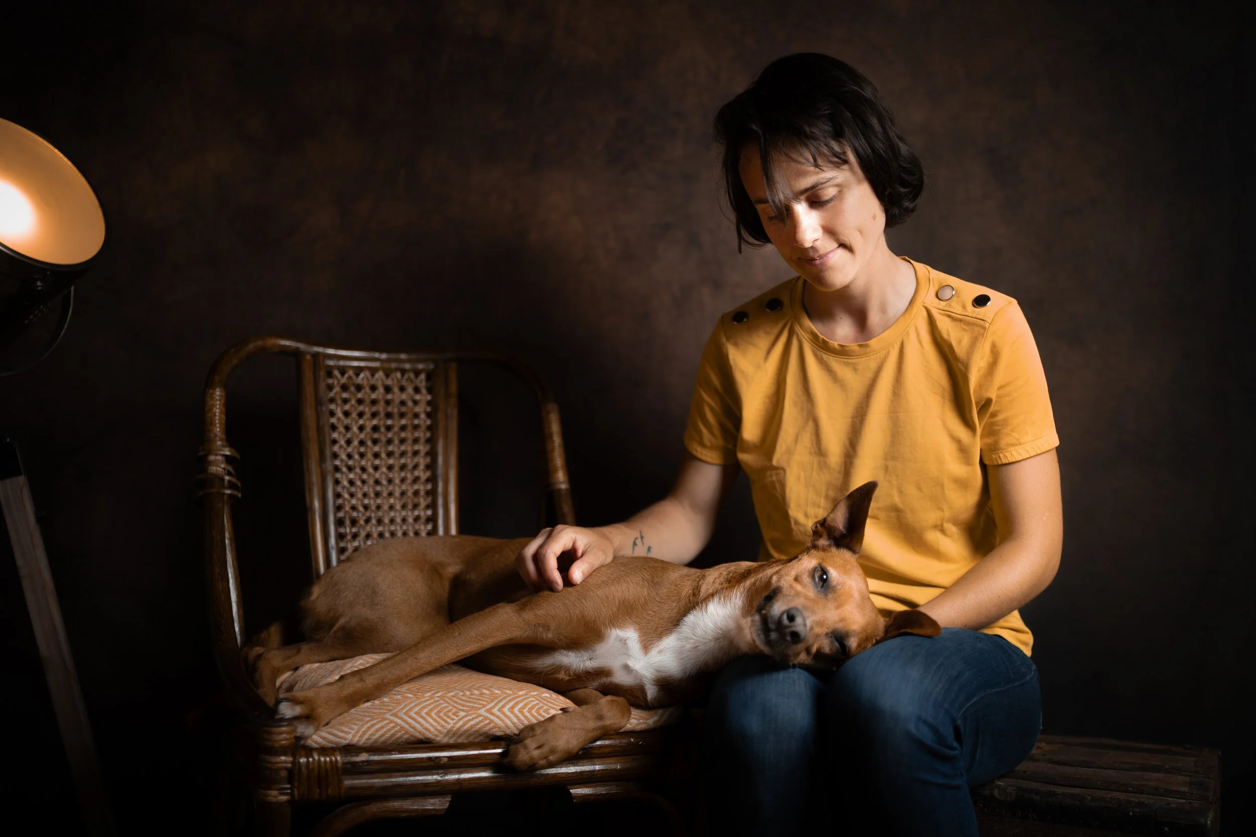 Une femme avec des cheveux noirs courts, portant un t-shirt jaune à épaulettes, caresse un chien allongé sur un fauteuil en osier. La scène est sombre avec un fond marron et une source de lumière chaude à gauche.