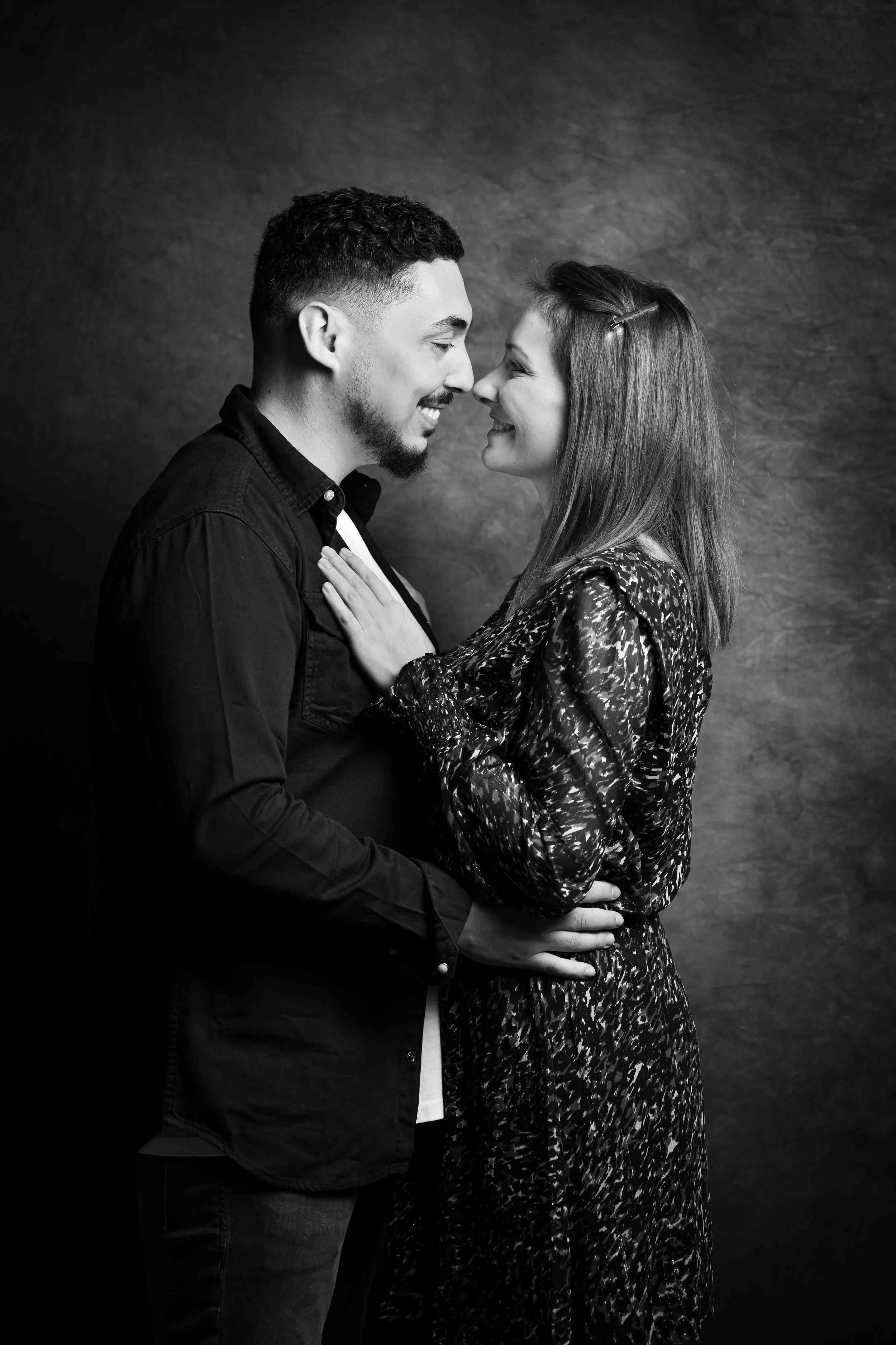 Un couple se tient face à face, se souriant, avec le front presque se touchant, dans une ambiance intime, en noir et blanc.