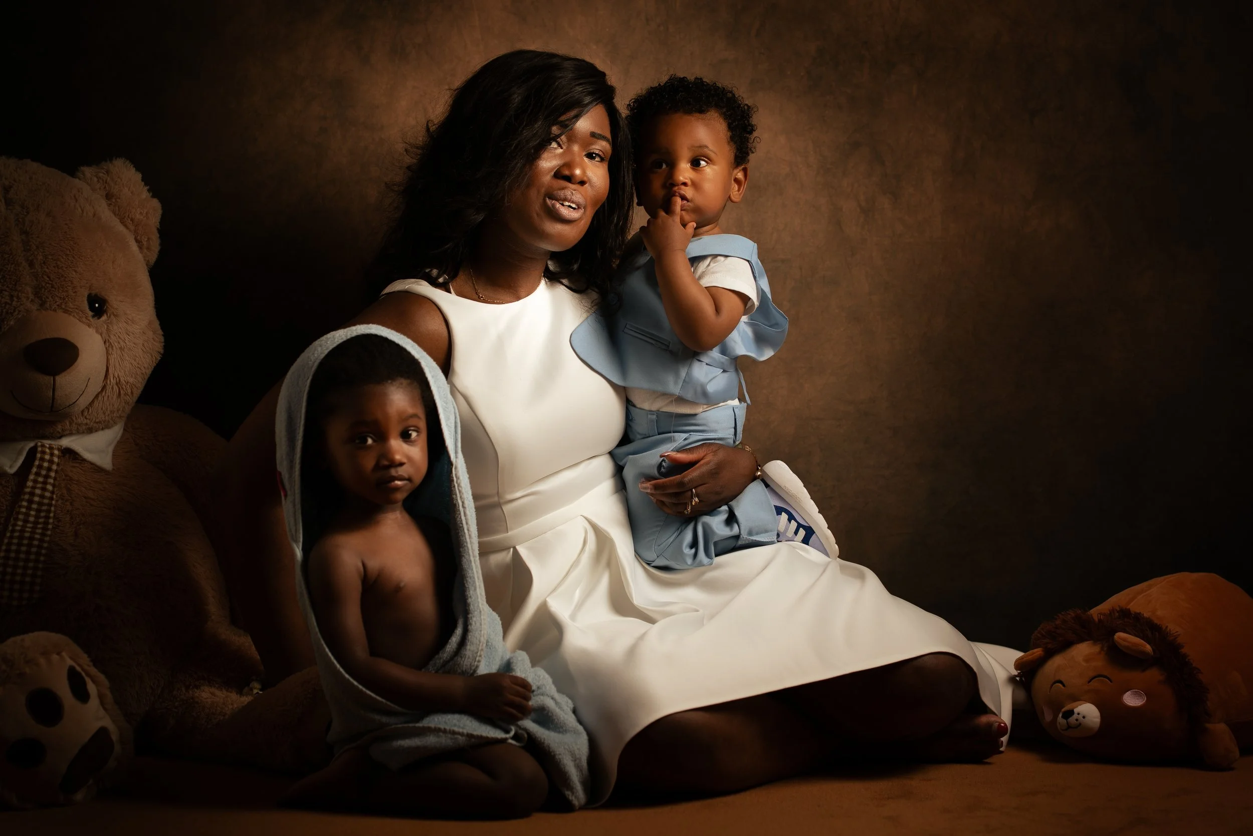Une femme noire avec deux enfants, un garçon et une fille, assis sur un canapé entourés de peluches, dans une ambiance chaleureuse et intime.