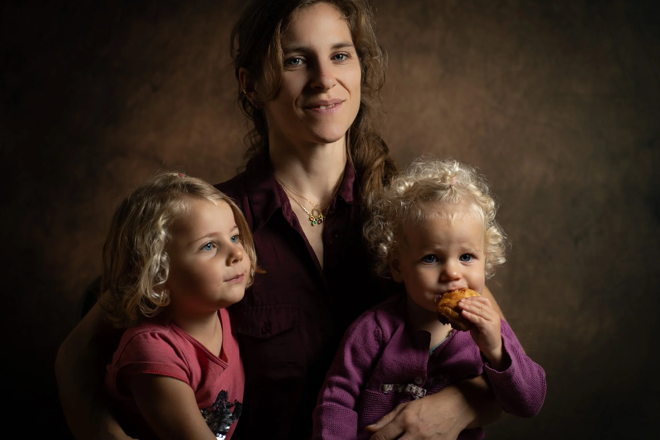 Une femme avec trois jeunes filles, deux blondes aux cheveux bouclés et une brune aux cheveux lisses, pose pour une photo dans un fond brun. La fille de droite mange un croissant et porte un pull violet. La femme porte un collier avec des pendentifs 