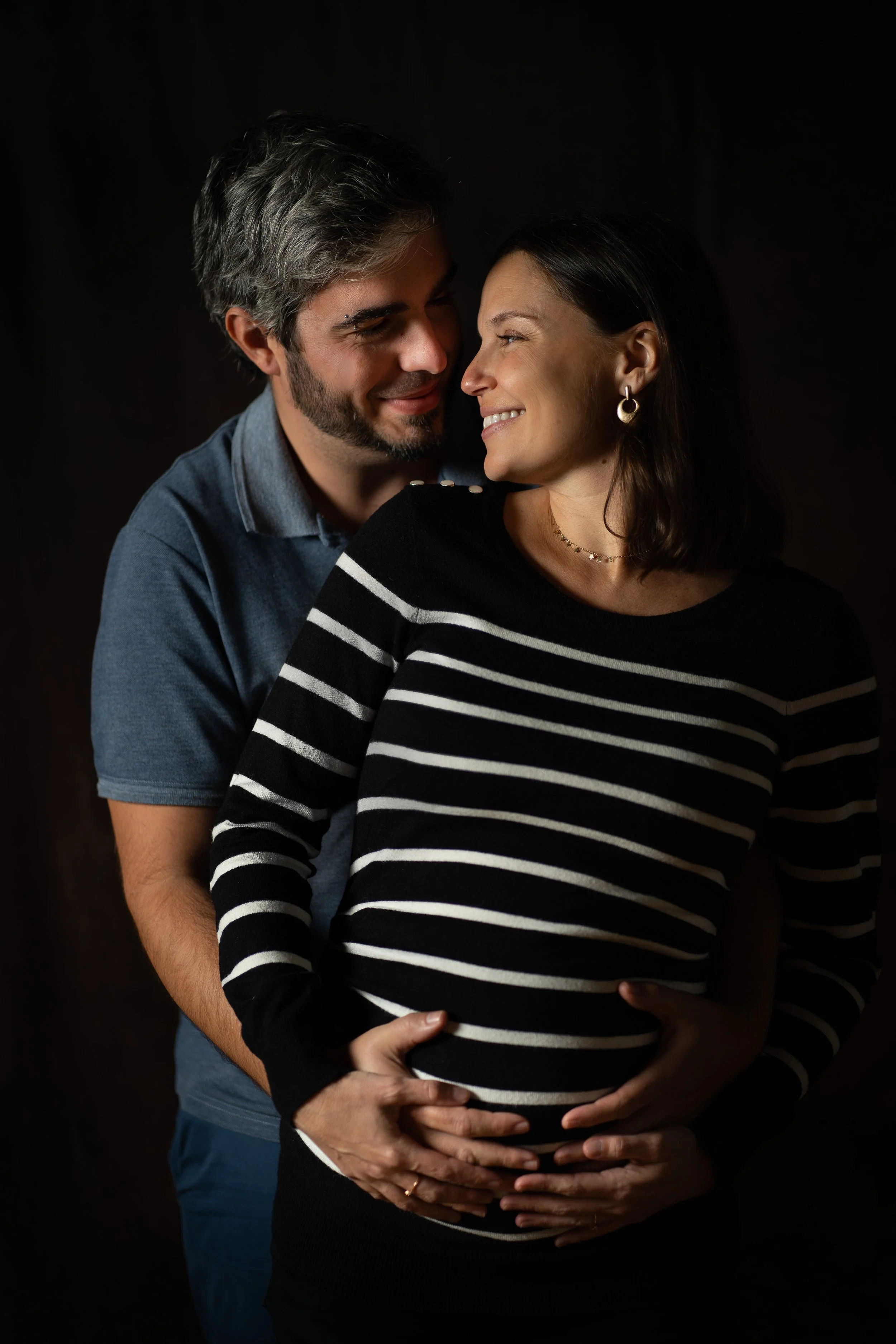 Un couple se tient derrière une femme enceinte, tous souriants. La femme porte un pull rayé noir et blanc et des boucles d'oreilles en forme de cercle.