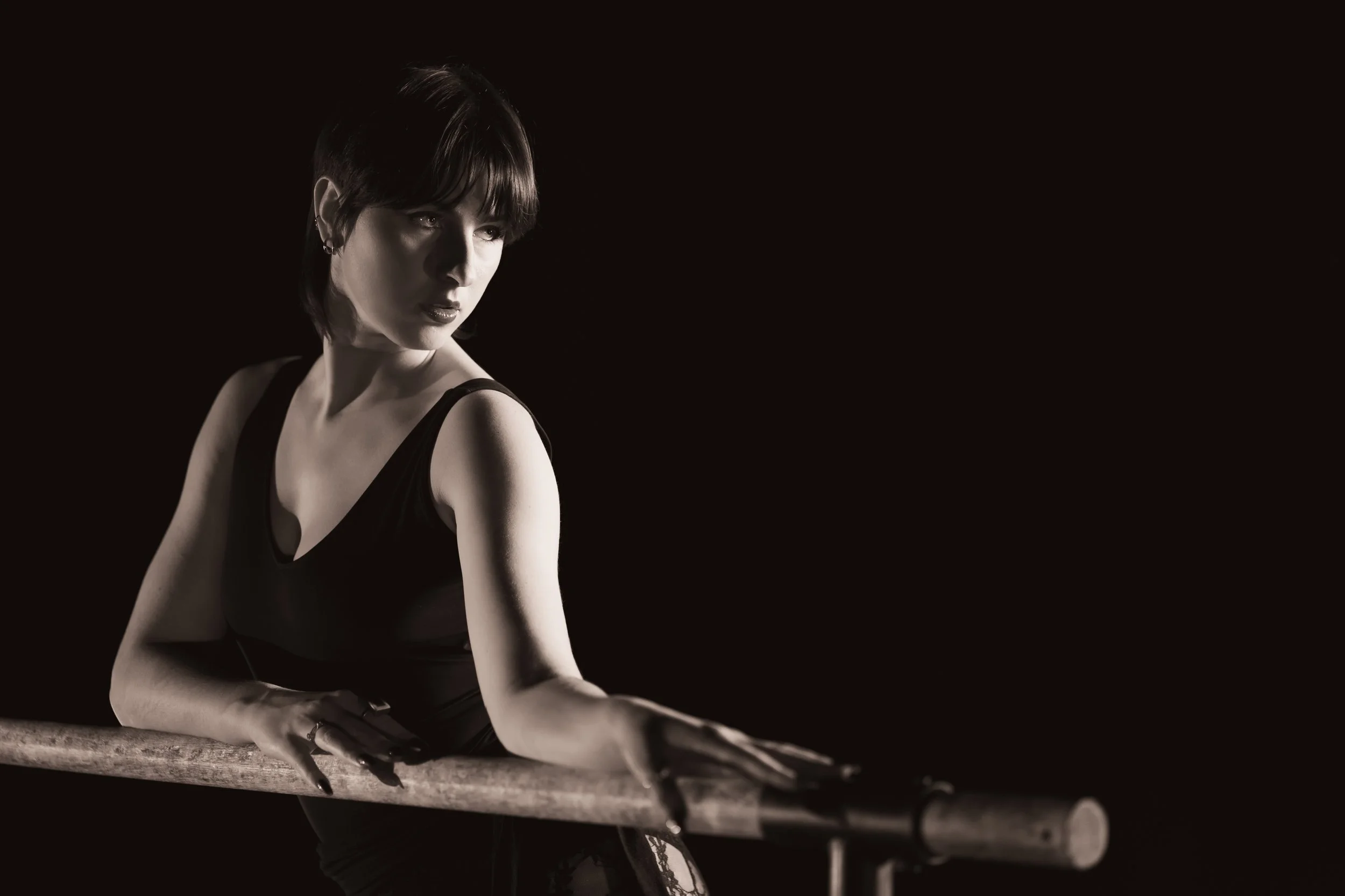 Une femme aux cheveux courts et foncés, portant un haut noir, posant sur une barre de ballet en latex noir, dans un environnement sombre avec un éclairage dramatique.