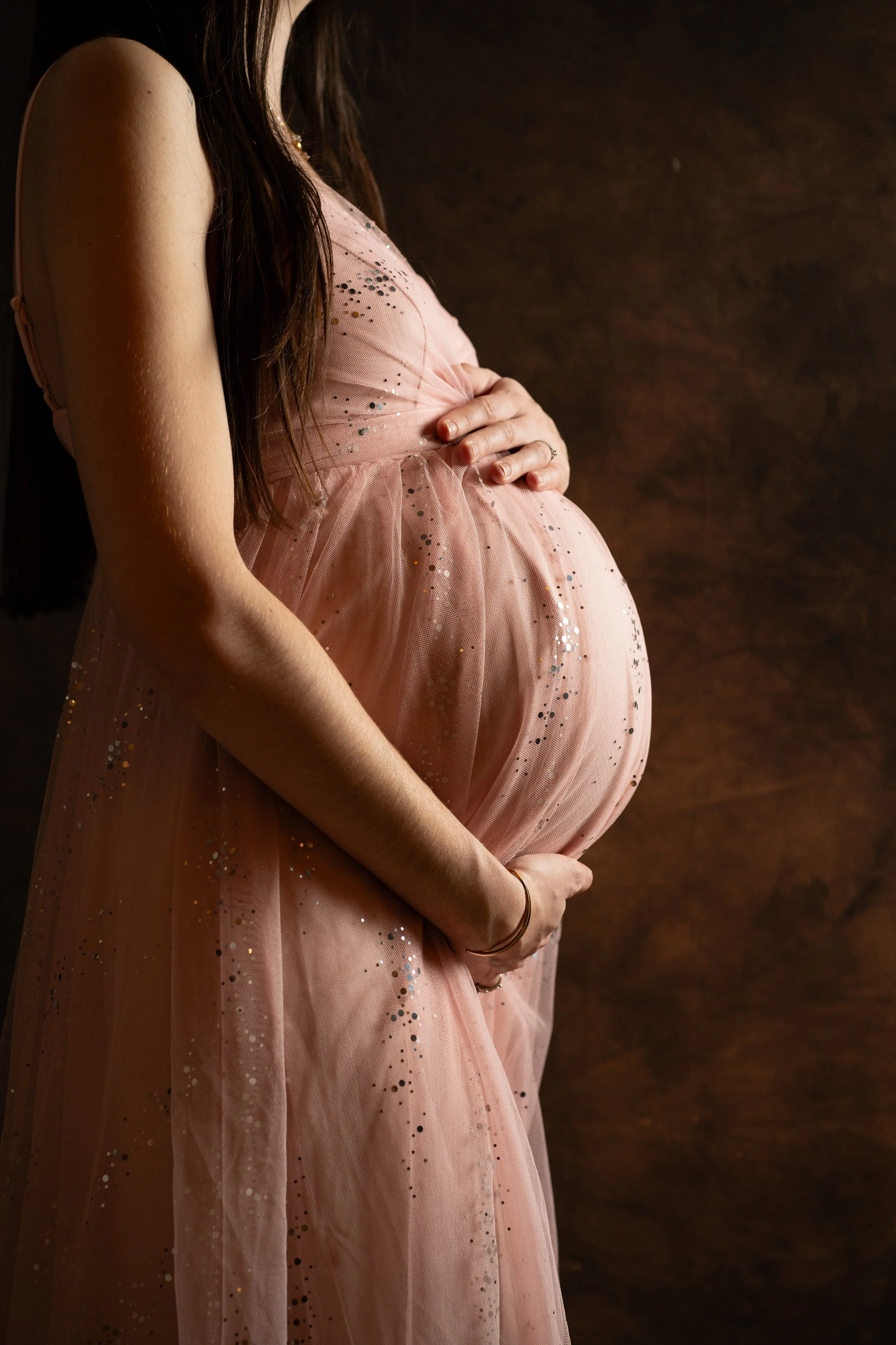 Femme enceinte portant une robe rose clair avec des paillettes, tenant son ventre, contre un fond sombre.