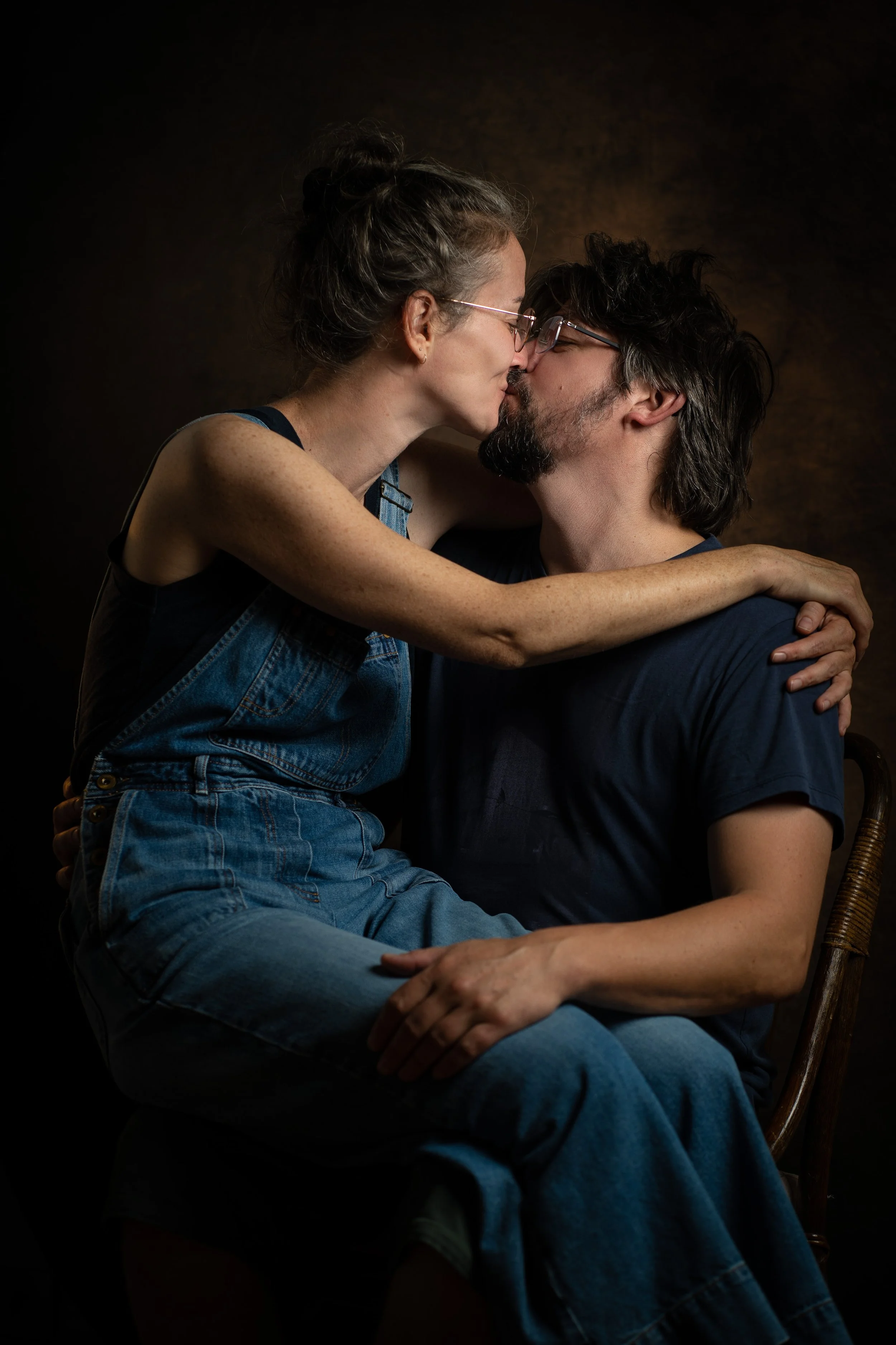 Un couple s'embrasse tendrement, la femme est assise sur les genoux de l'homme, avec un fond sombre.