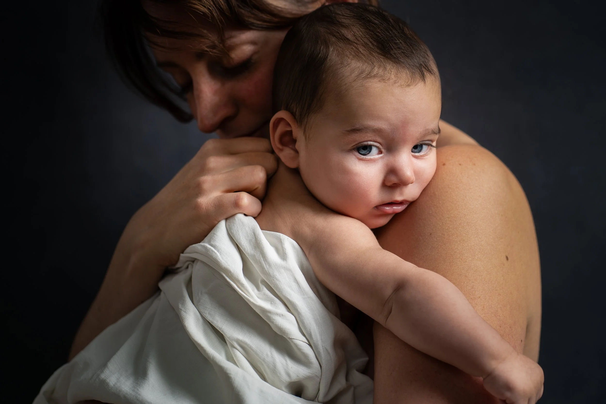 Un adulte tient un jeune garçon contre lui dans une posture protectrice, le visage du garçon est visible avec des yeux bleus et un regard concentré. La scène est sombre avec un fond noir.