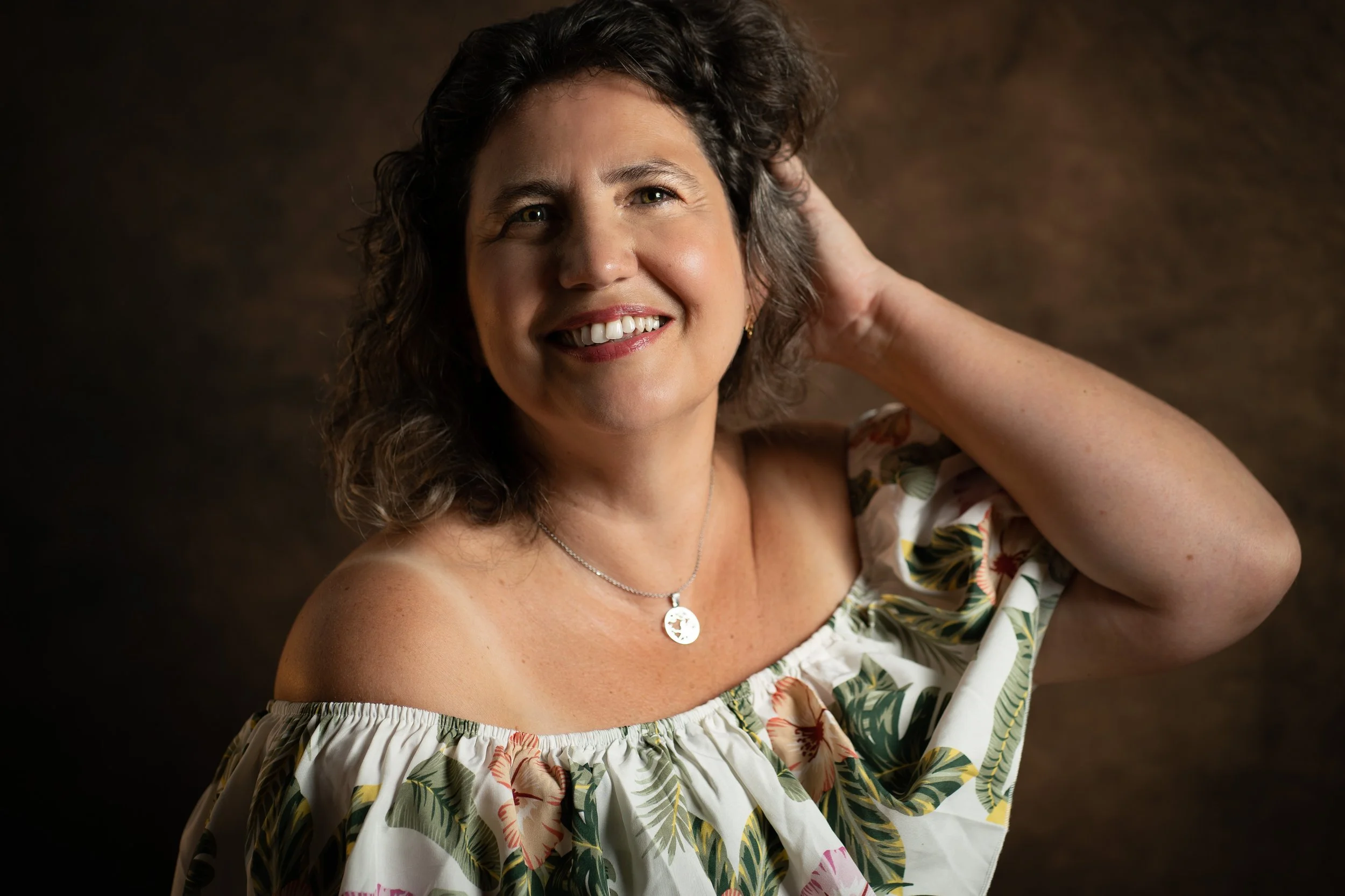 Une femme souriante avec des cheveux bruns, portant un collier en argent et une robe à motifs floraux. Elle pose avec une main derrière la tête contre un fond sombre.