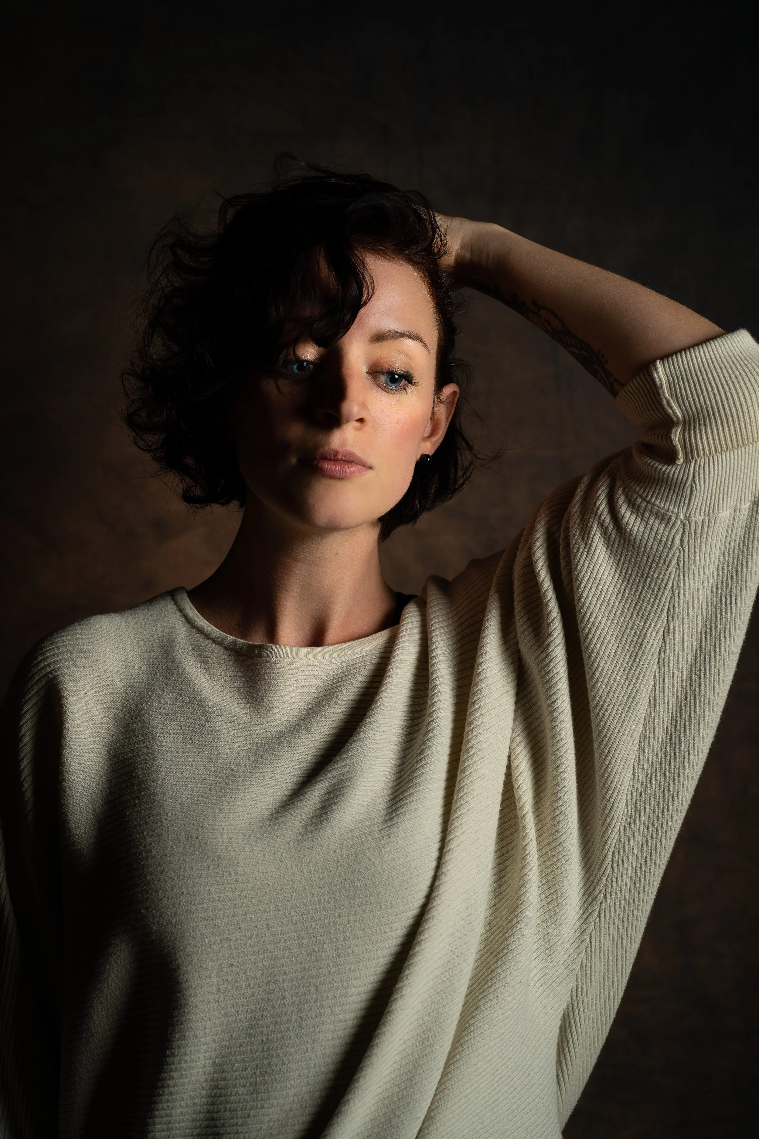 Jeune femme avec cheveux courts et bouclés, portant une veste en tricot beige, avec un tatouage sur le bras, regardant légèrement à gauche, fond sombre.