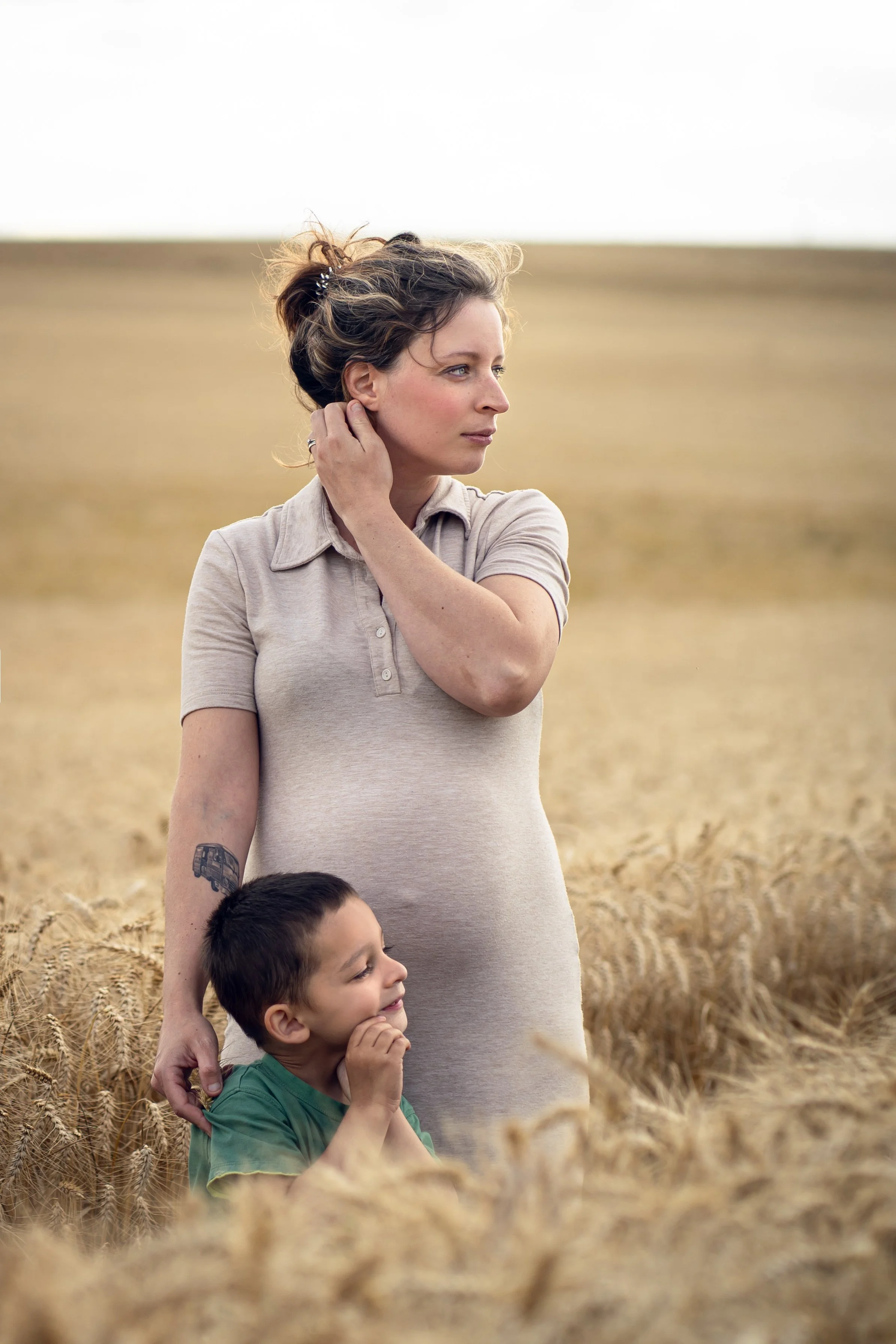 Une femme et deux enfants se tiennent dans un champ de blé, regardant vers la droite. La femme a les cheveux attachés en chignon, et l’un des enfants la serre contre elle. Le paysage est en plein air avec un ciel clair.