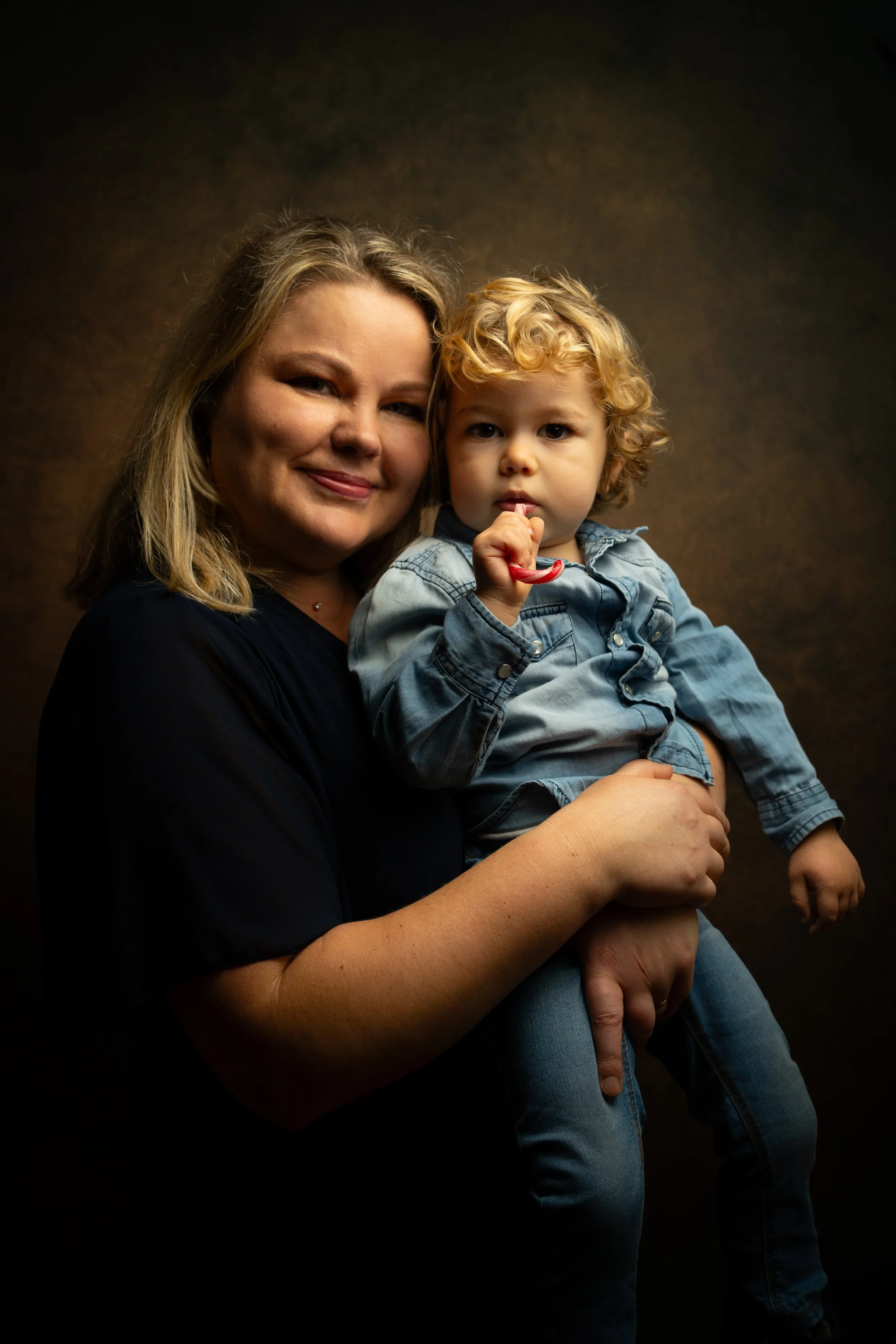 Une femme souriante tient une petite fille aux cheveux bouclés, qui a un mouchoir dans la bouche, dans un studio avec un fond sombre.