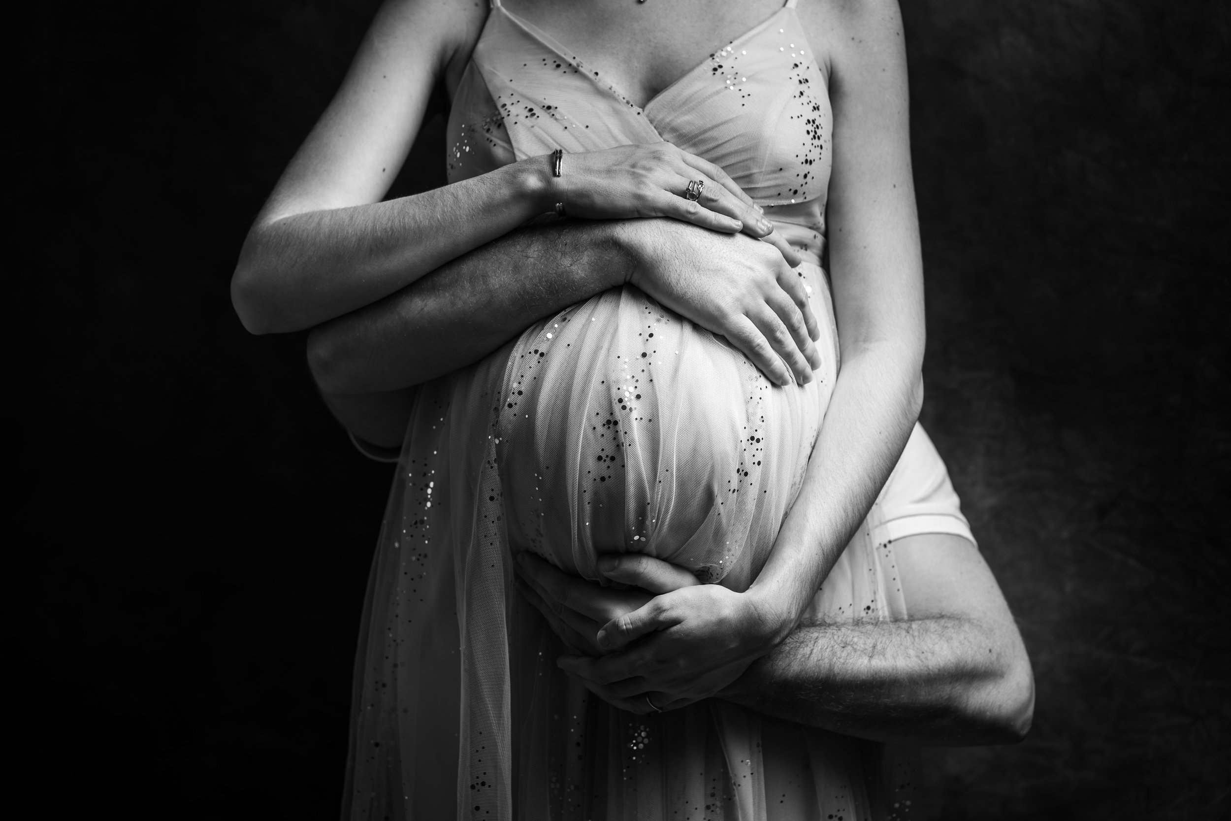 Une femme enceinte portant une robe élégante, étant soutenue par un homme dont on ne voit que les bras, dans une composition en noir et blanc.