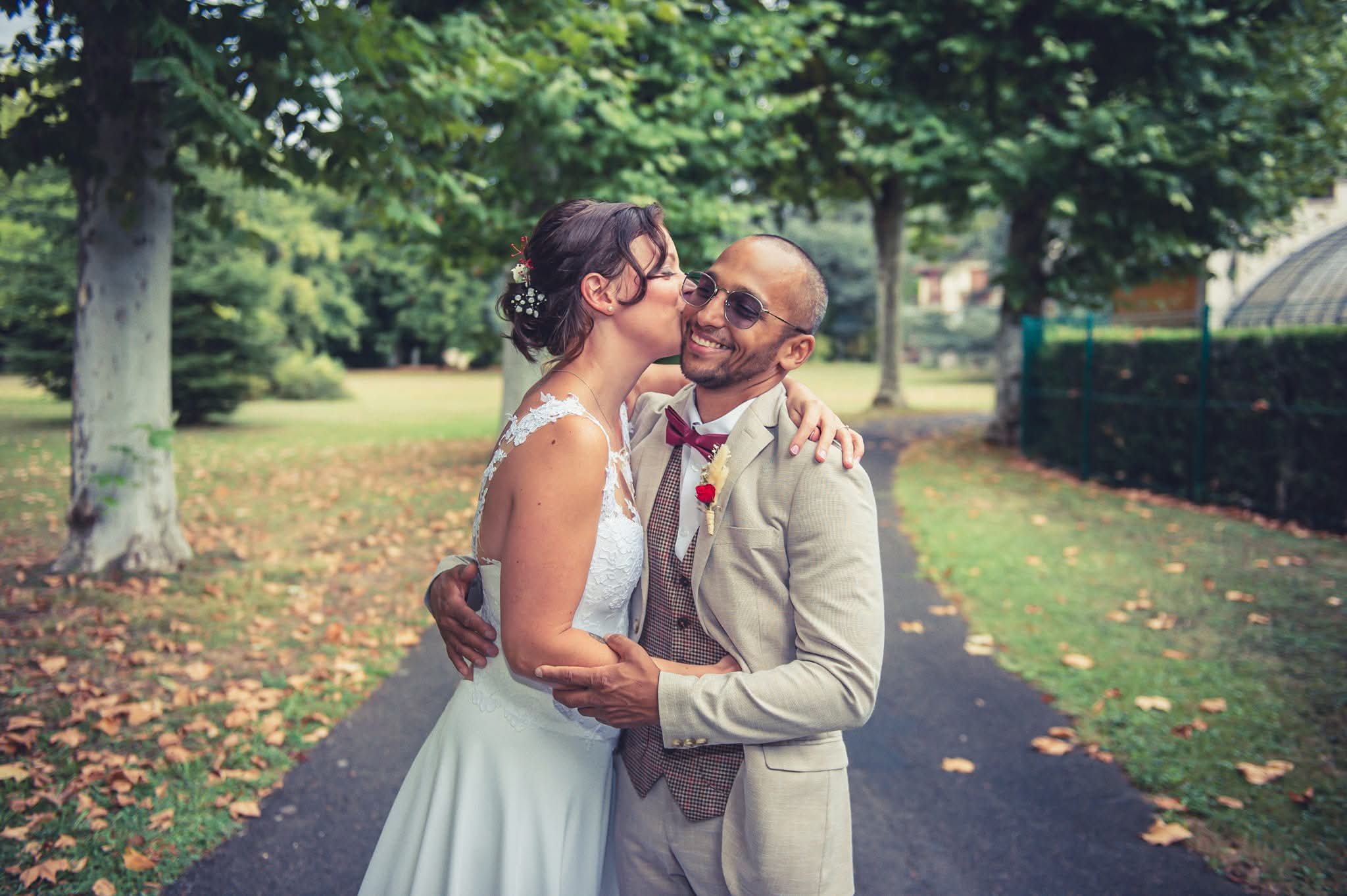 Un couple de mariés échange un baiser dans un parc verdoyant avec des arbres et des feuilles sur le sol.