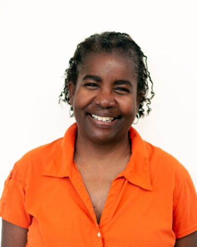 A smiling woman with curly hair wearing an orange shirt against a white background.