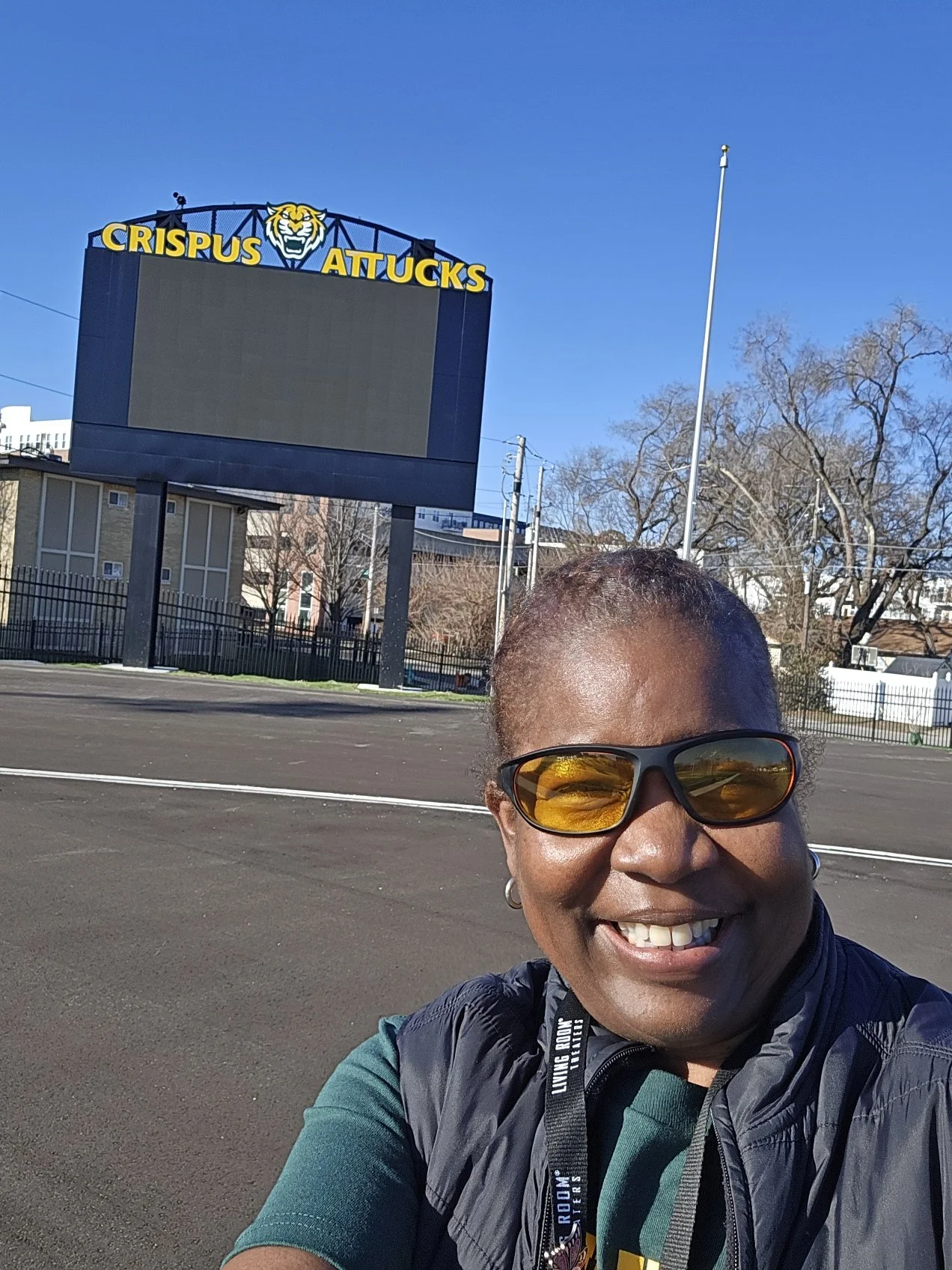 A woman smiling and taking a selfie outdoors with a blue clear sky, bare trees, and an empty sports field visible in the background. A large digital signboard with a tiger logo and the text 'CRISPUS ATTUCKS' is also in the background.