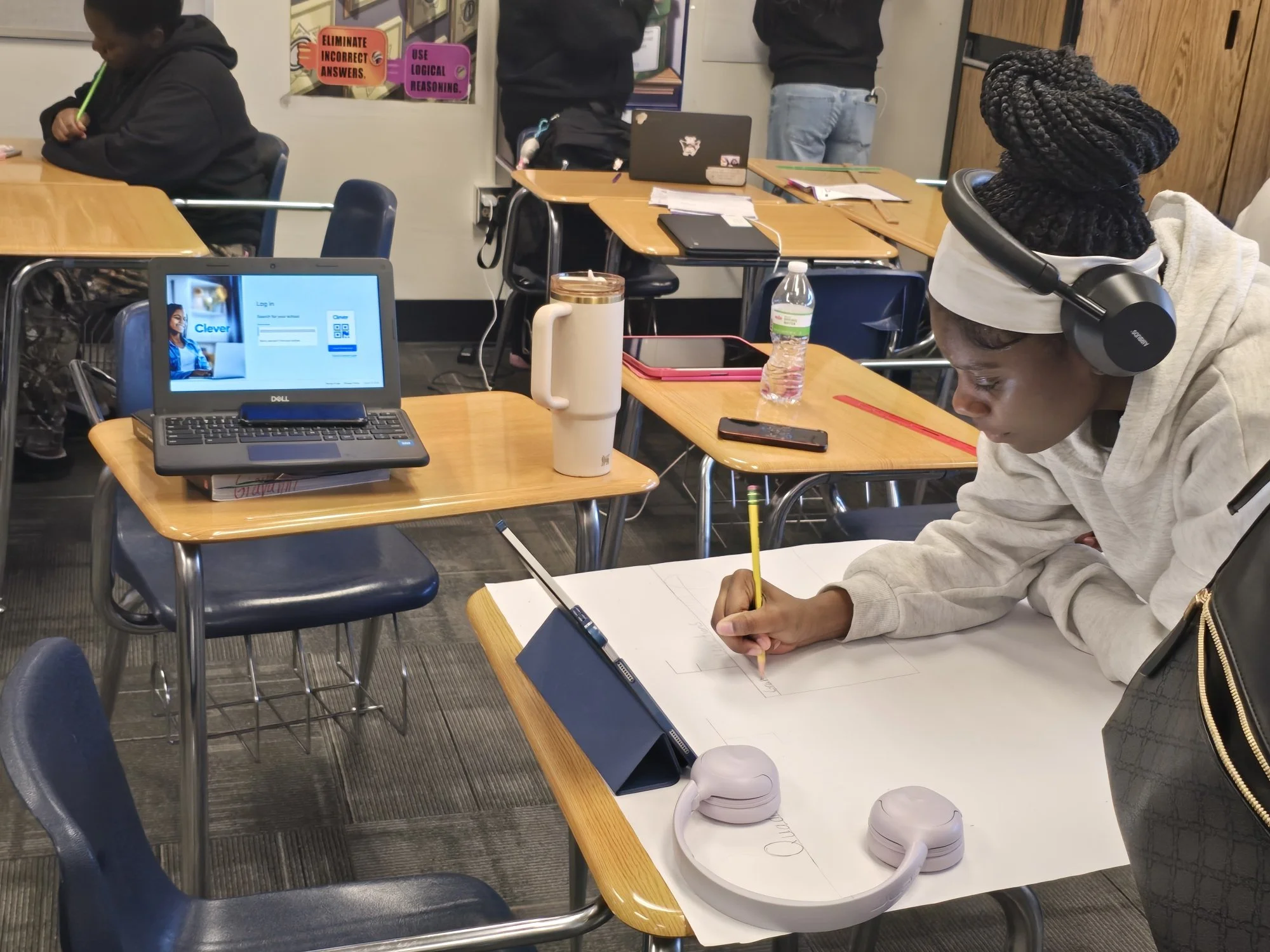 A classroom with students seated at desks, some working and others using laptops or wearing headphones. One student in the foreground is drawing or writing on paper, wearing headphones and a hooded sweatshirt. There are water bottles and notebooks on the desks, and the background shows classroom posters and other students.