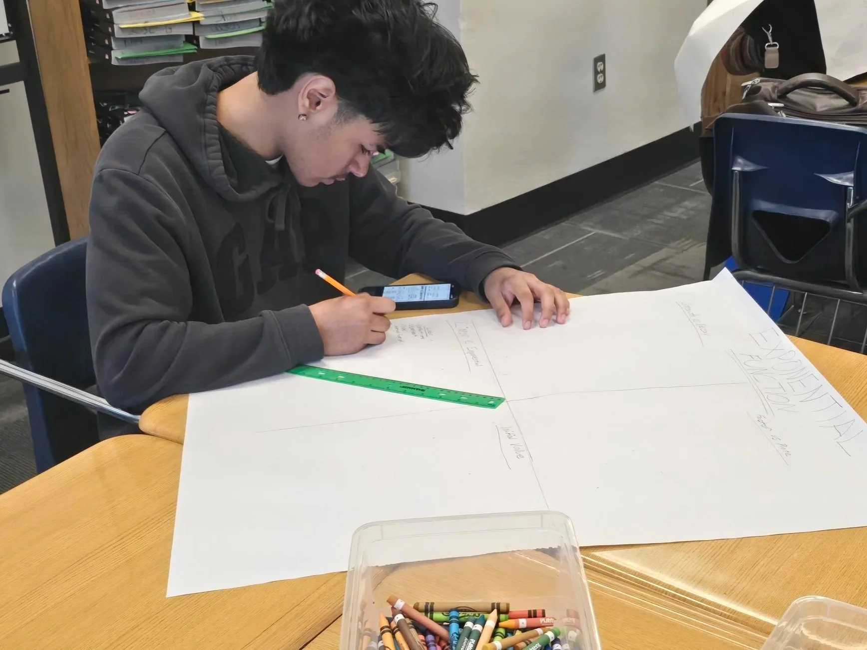 A young man with dark hair and earrings sitting at a table, writing on a large sheet of paper with a pencil. He is wearing a dark hoodie. There is a ruler on the paper, and a smartphone placed beside his hand. The table has some color crayons in a plastic container. In the background, there are shelves with stacked files or folders, and a chair with a bag on it.