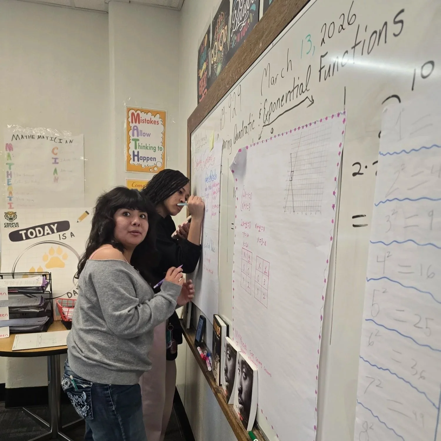 Two women are working at a whiteboard filled with notes and charts in a classroom; one woman is writing with a marker while the other looks at the camera.