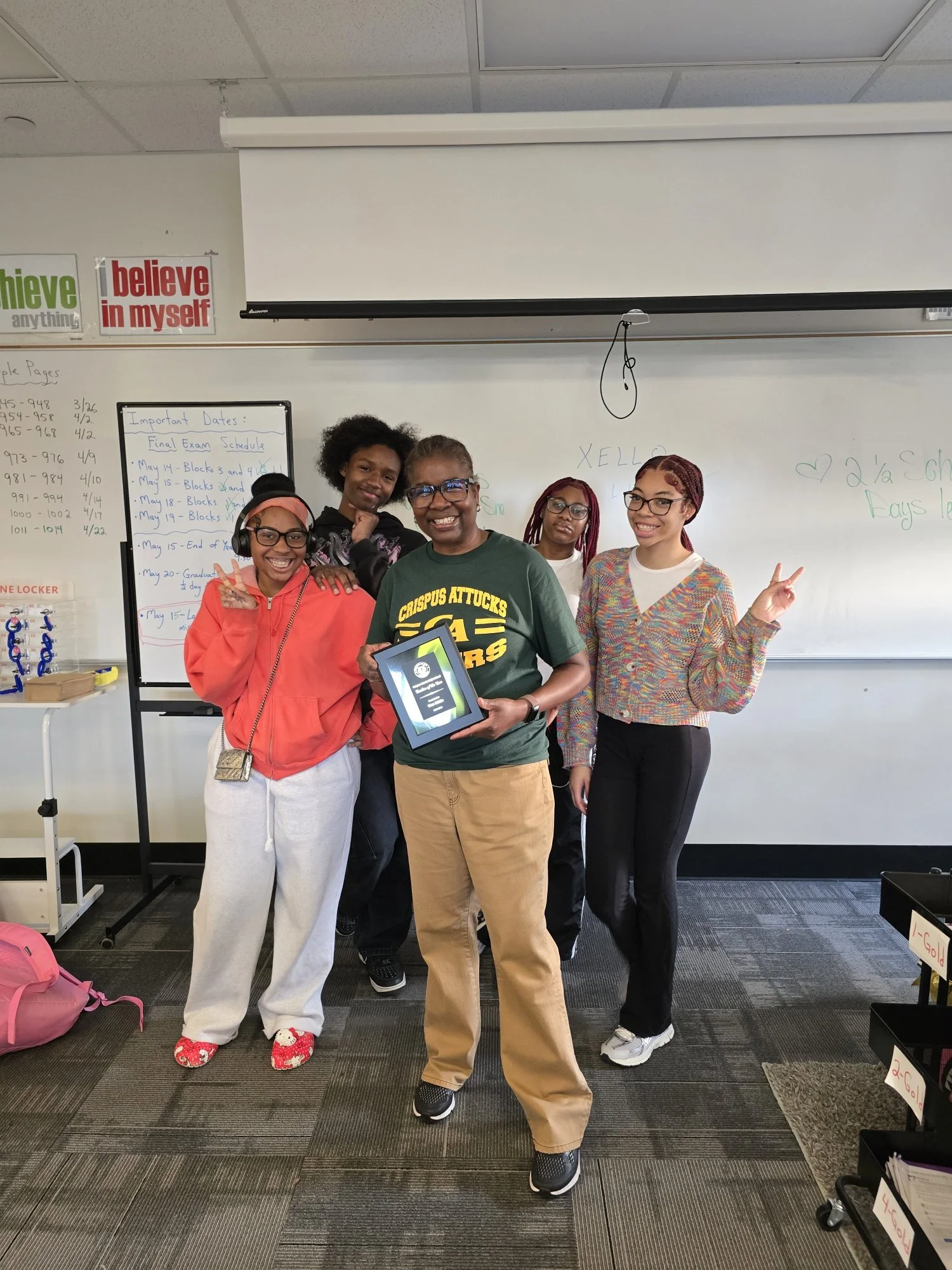 A group of five women standing together in a classroom, smiling and posing for a photo, some making peace signs. One woman in front is holding a certificate.