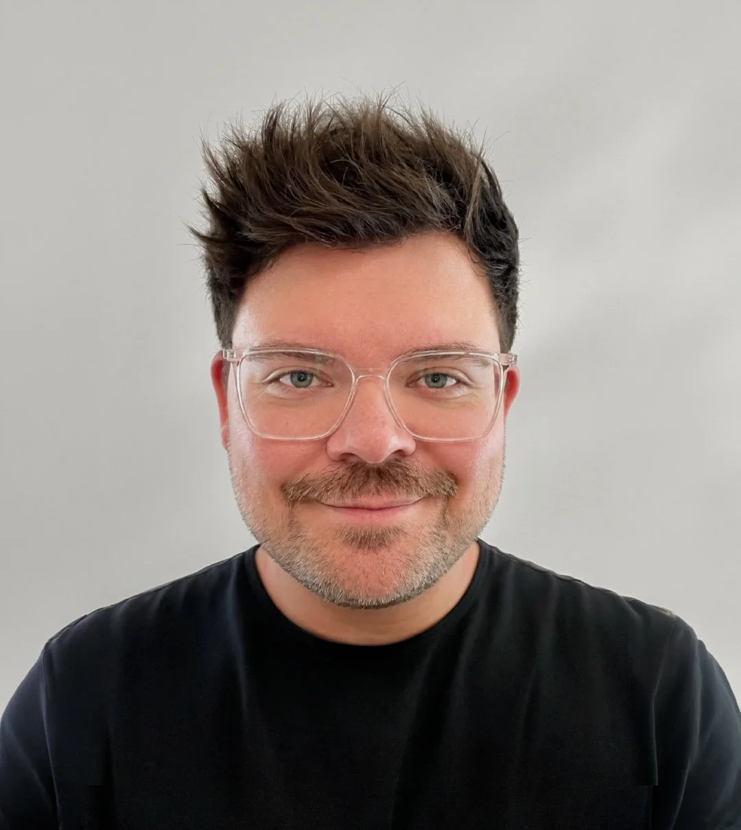Portrait of a smiling man with glasses and dark hair, wearing a black shirt, against a plain light background.