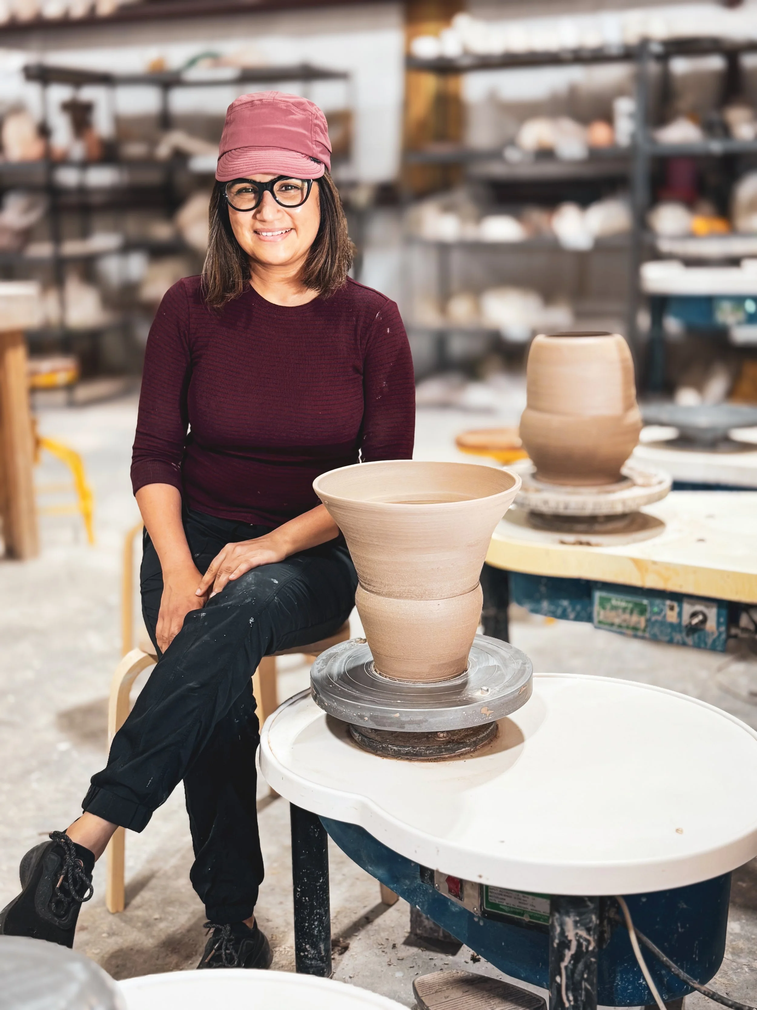 A woman with glasses and a pink hat sitting beside a pottery wheel in a studio with shelves of pottery in the background. She is smiling and wearing a maroon top and black pants, with a partially finished large clay vase on the wheel in front of her.