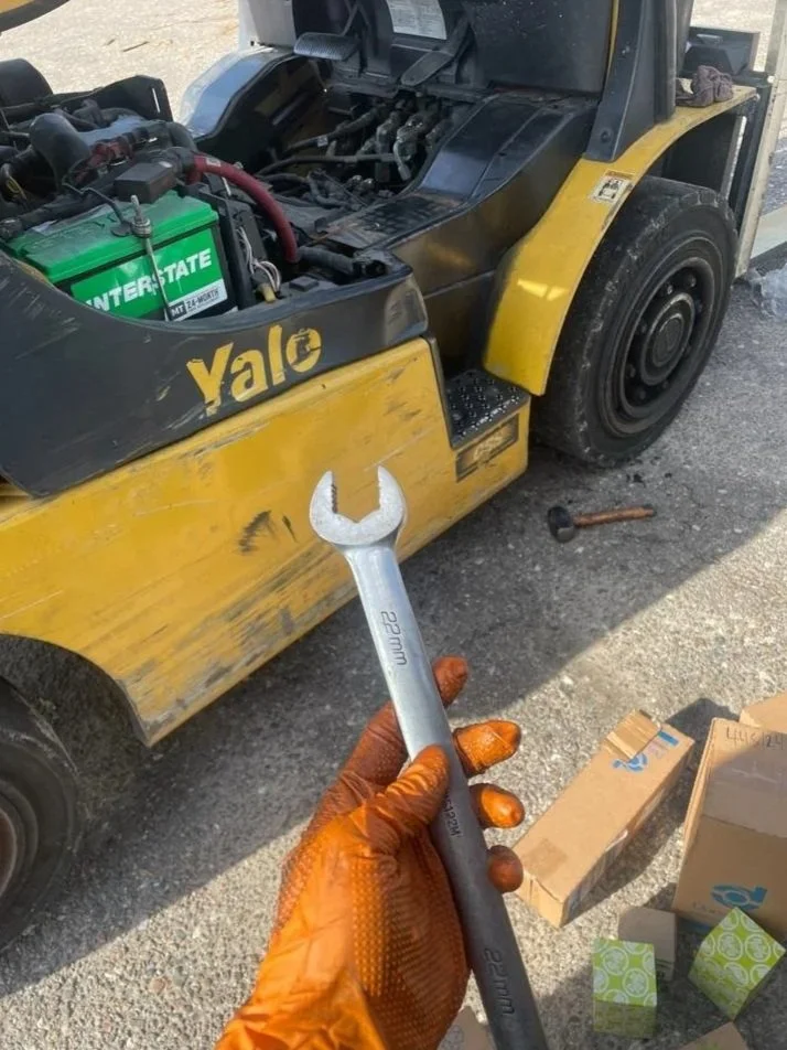 Close-up of a wrench held by a person wearing orange gloves, pointing at a yellow forklift with the hood open, showing the battery and internal components, on a gravel surface.
