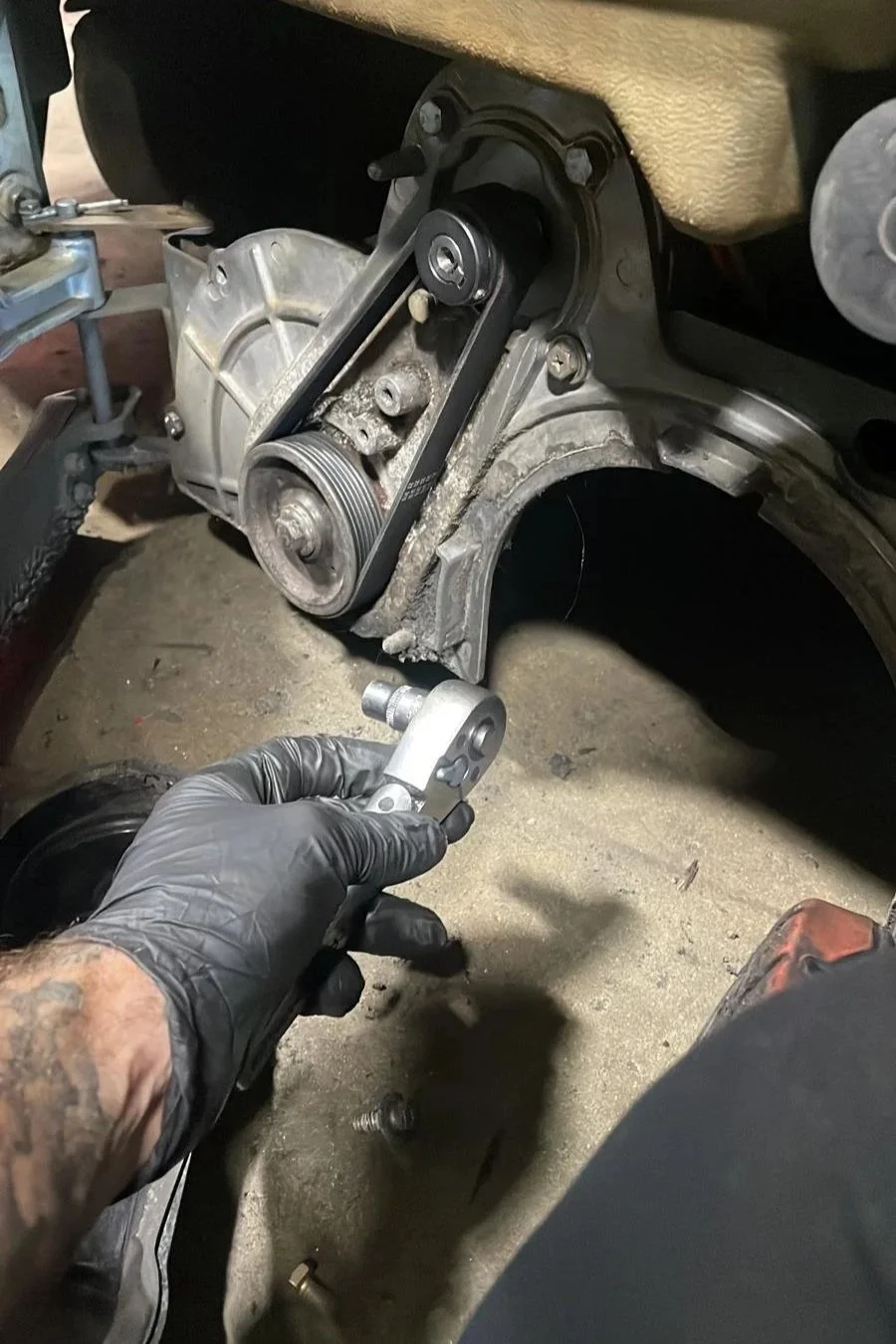 A person wearing black gloves holding a tensioner tool near an automotive engine component, with a belt installed on a pulley system and an engine block in the background.