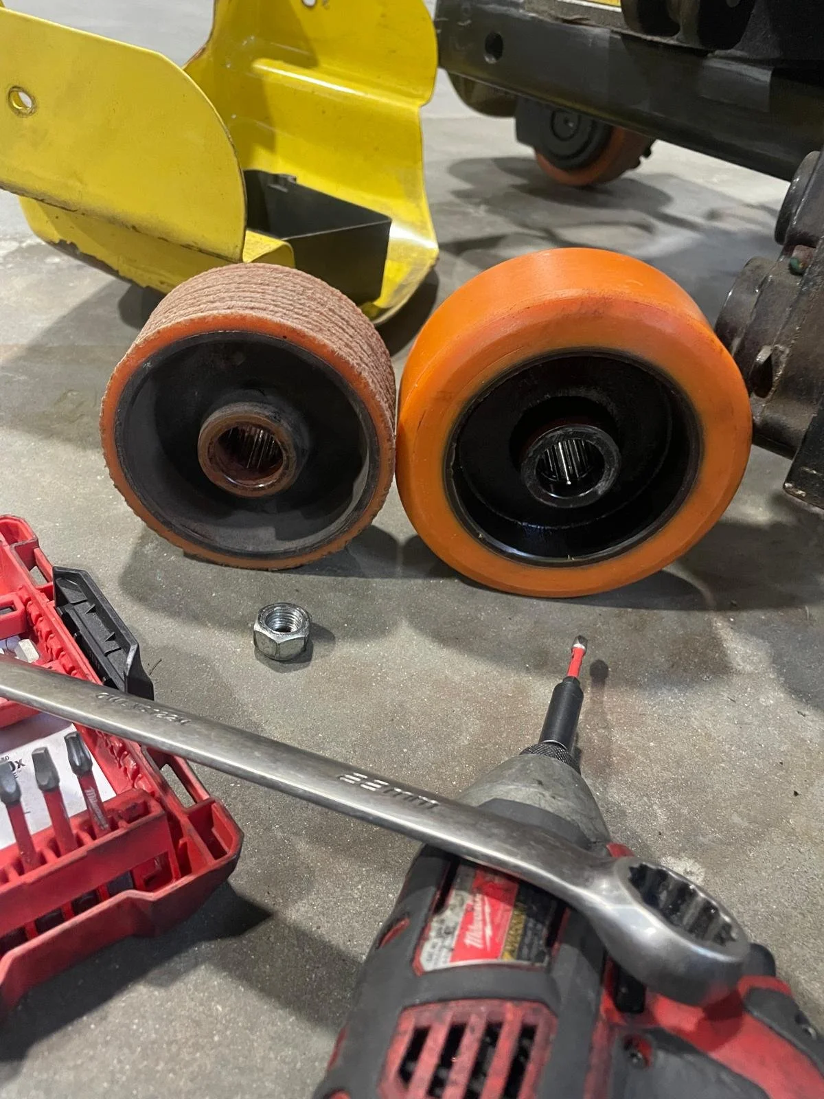 Two replacement forklift wheels, one worn and one new, near a power tool, nuts, and tools on a concrete surface, with a yellow object and part of a metal structure in the background.