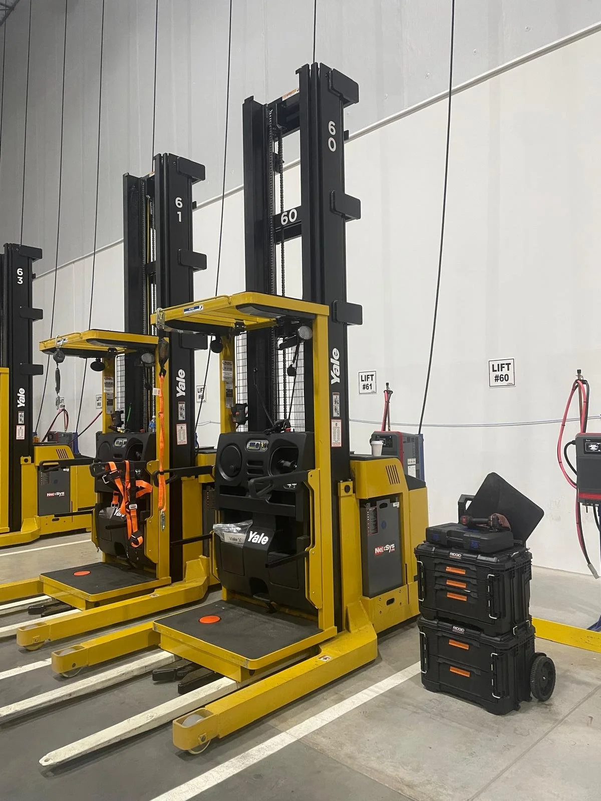 Multiple yellow Yale electric forklifts lined up in a warehouse. Some forklifts have safety harnesses hanging. There are black tool cases and equipment on the ground beside them. The warehouse wall is plain with signs indicating lift numbers.