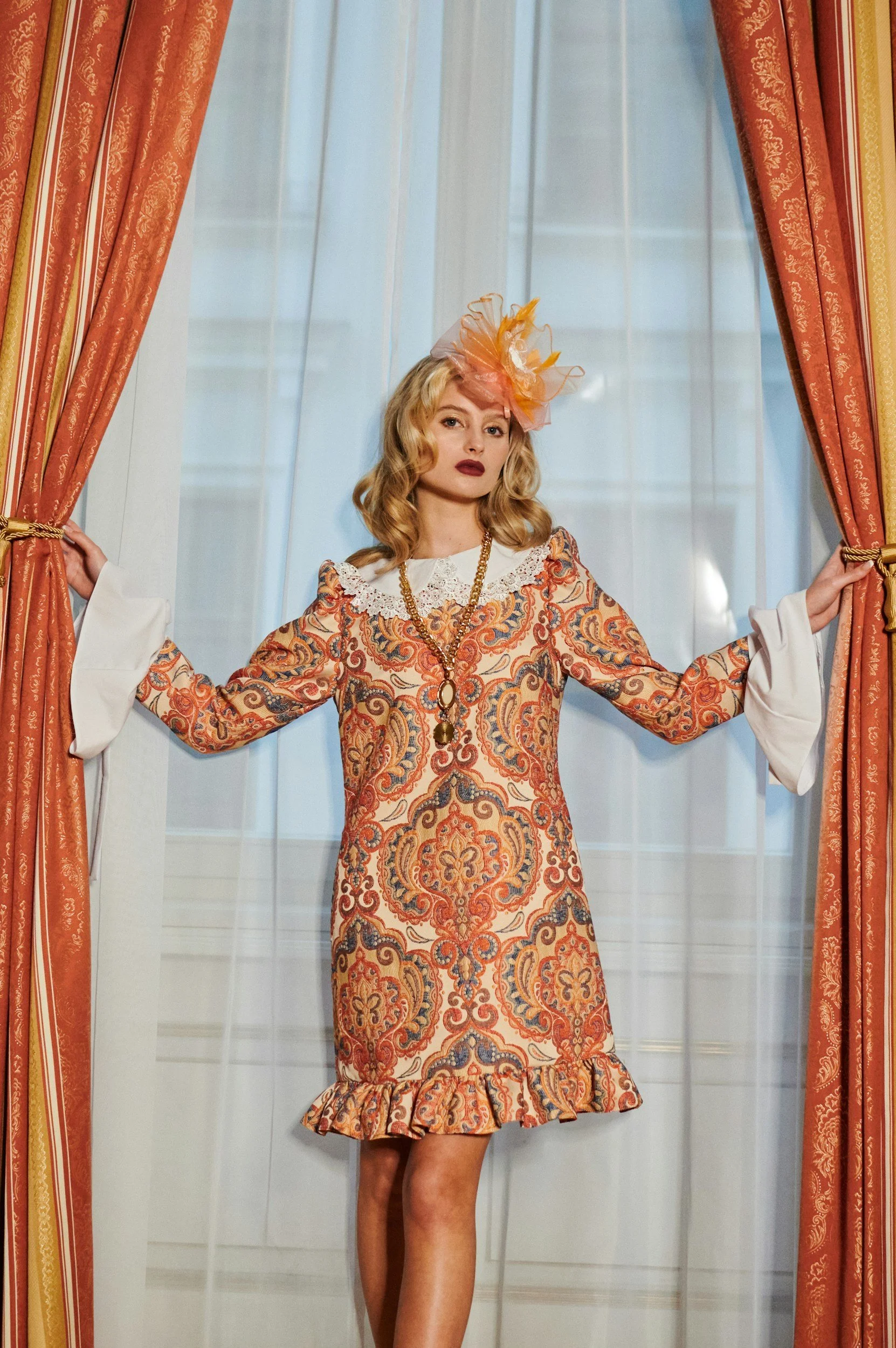 A woman with blonde hair wearing a patterned dress with ruffle hem, a large orange fascinator, and gold jewelry, standing behind orange curtains with a semi-transparent white curtain in the background.