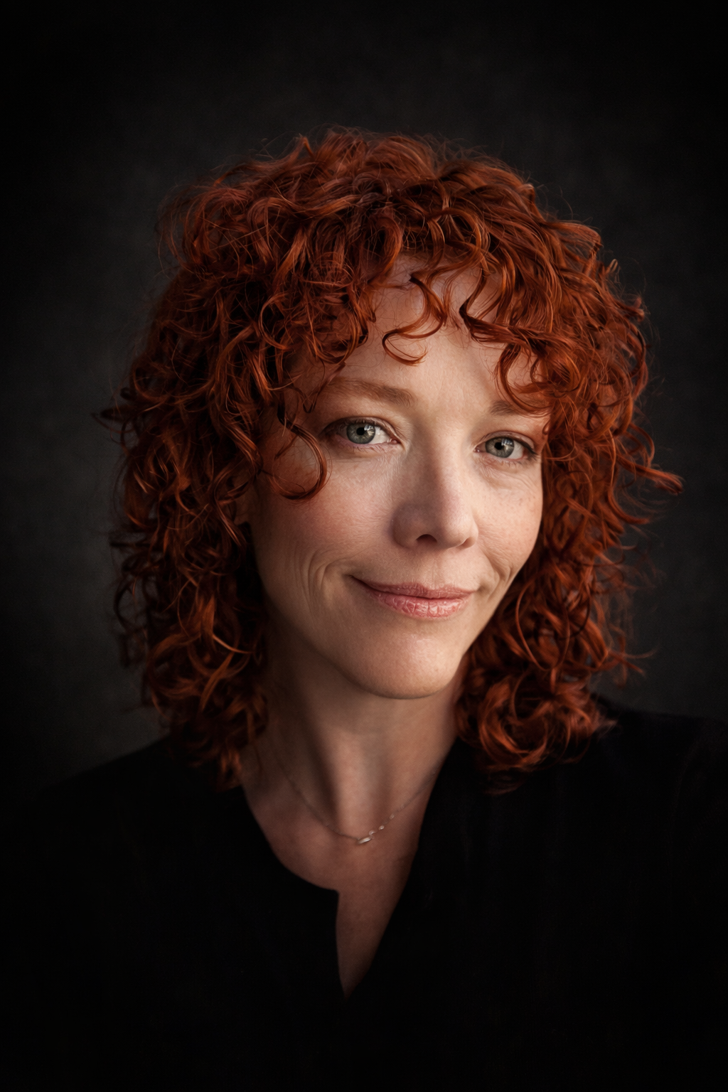 Close-up portrait of a woman with curly red hair, fair skin, and green eyes, wearing a black top and a delicate necklace, against a dark background.