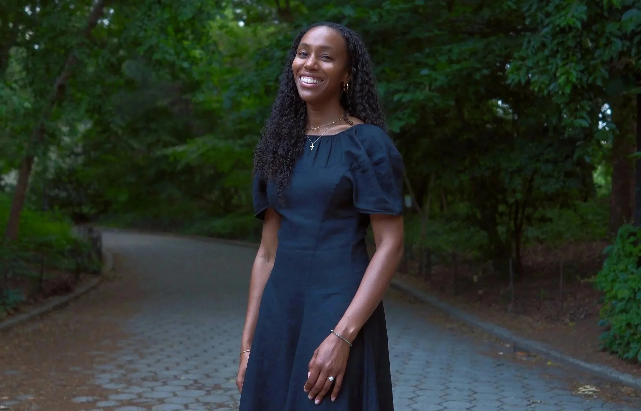 A woman with long curly hair wearing a black dress and jewelry, standing outdoors on a paved path surrounded by green trees, smiling at the camera.