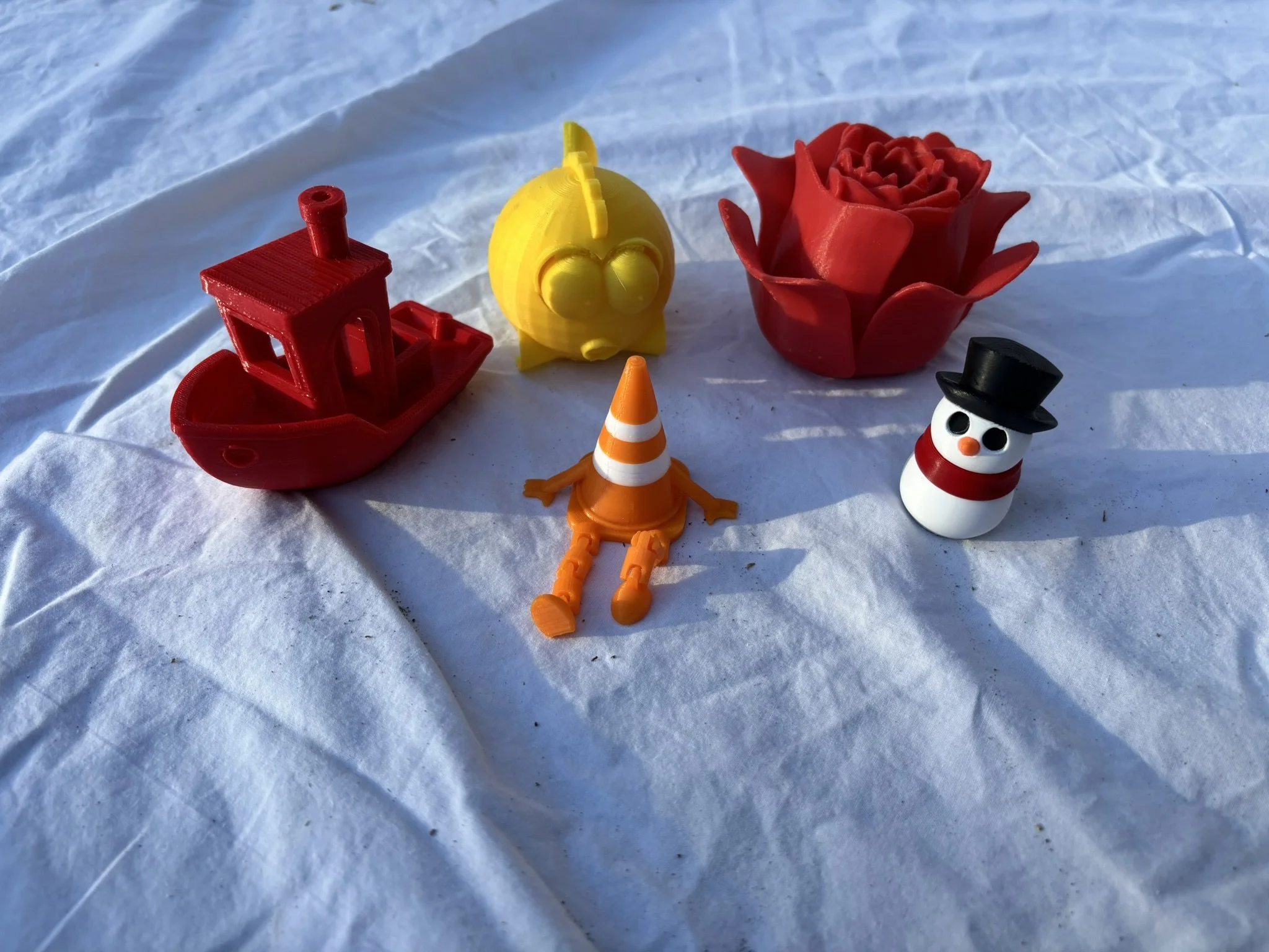 Colorful plastic toys arranged on a white surface, including a red boat, a yellow bird, a red rose, an orange traffic cone, and a small snowman with a black hat.