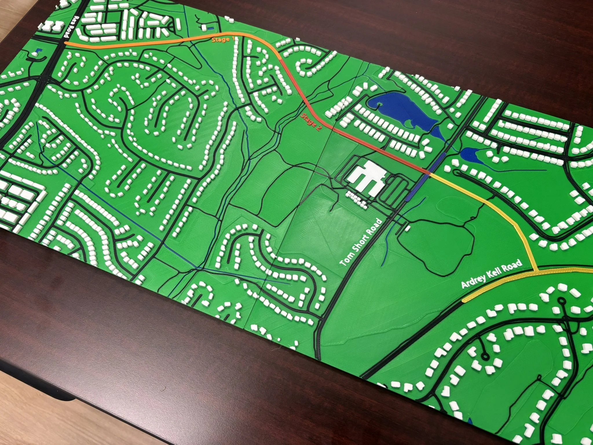 A detailed model of a suburban neighborhood map showing roads, residential areas, lakes, and stages labeled as 'Stage 1' and 'Stage 2' on a wooden table.