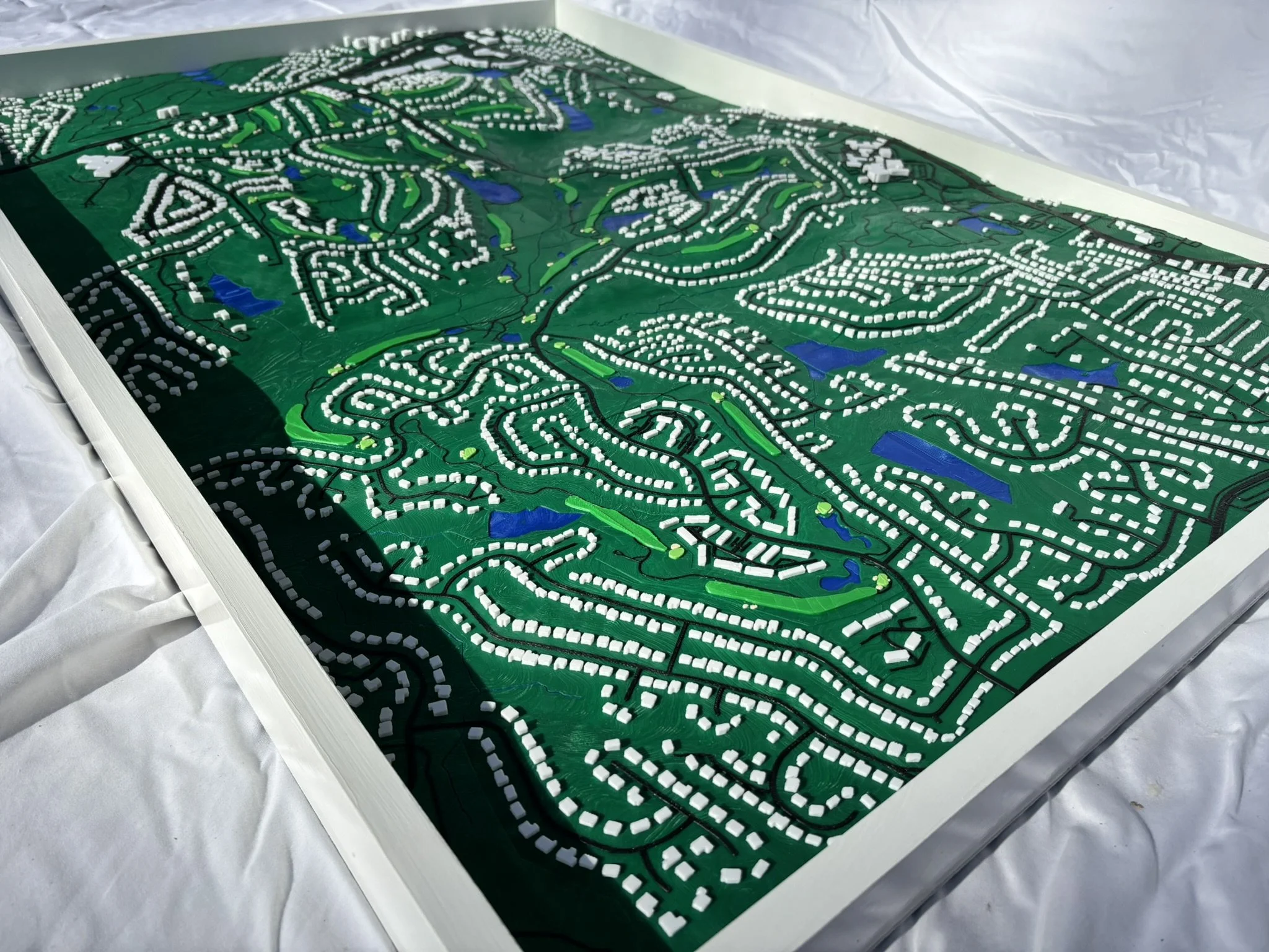 3D topographical map model with green terrain, water features, white structures, and black lines representing roads or pathways, placed on a white surface.
