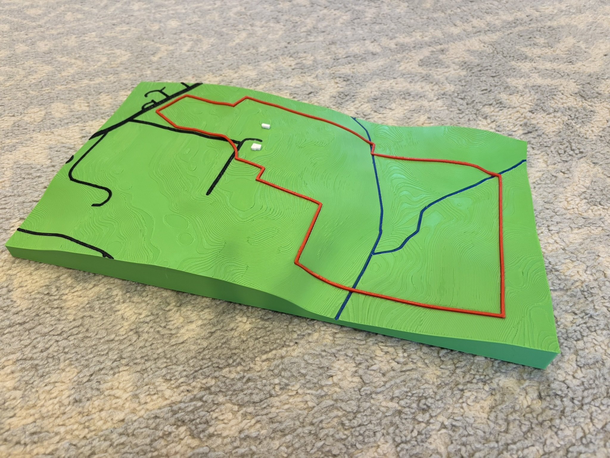 A detailed topographic map on a green surface showing contour lines, with colored lines and two small white structures.