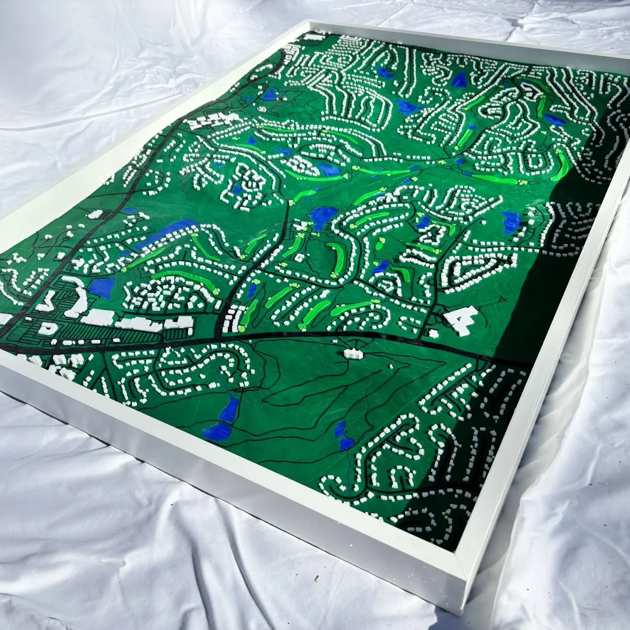 A detailed scale model of a suburban neighborhood with winding roads, green spaces, and small blue water bodies, framed in white and resting on a white surface.