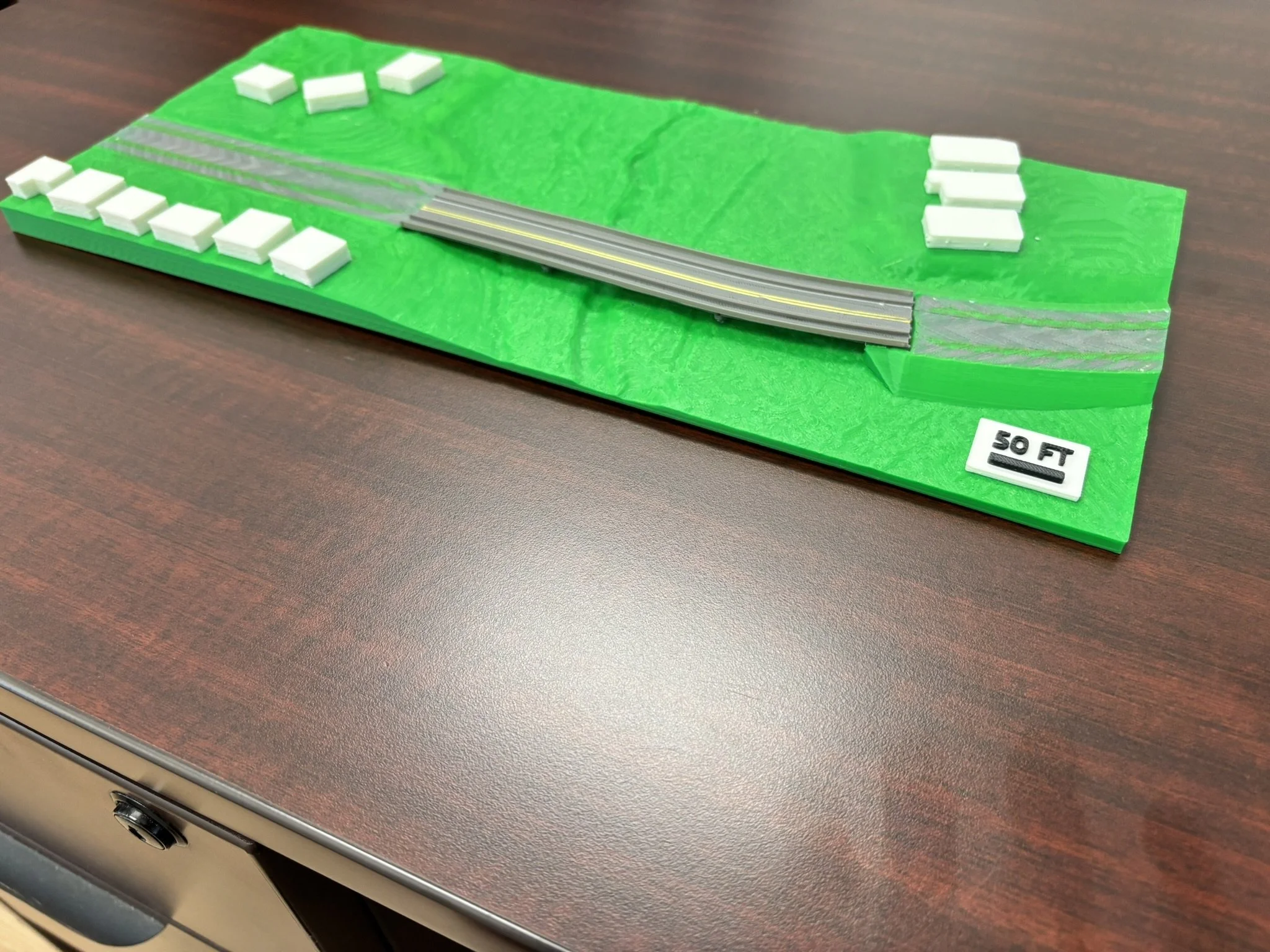 A  model of a landscape with a road, bridge, grass, and small white structures, placed on a wooden surface.