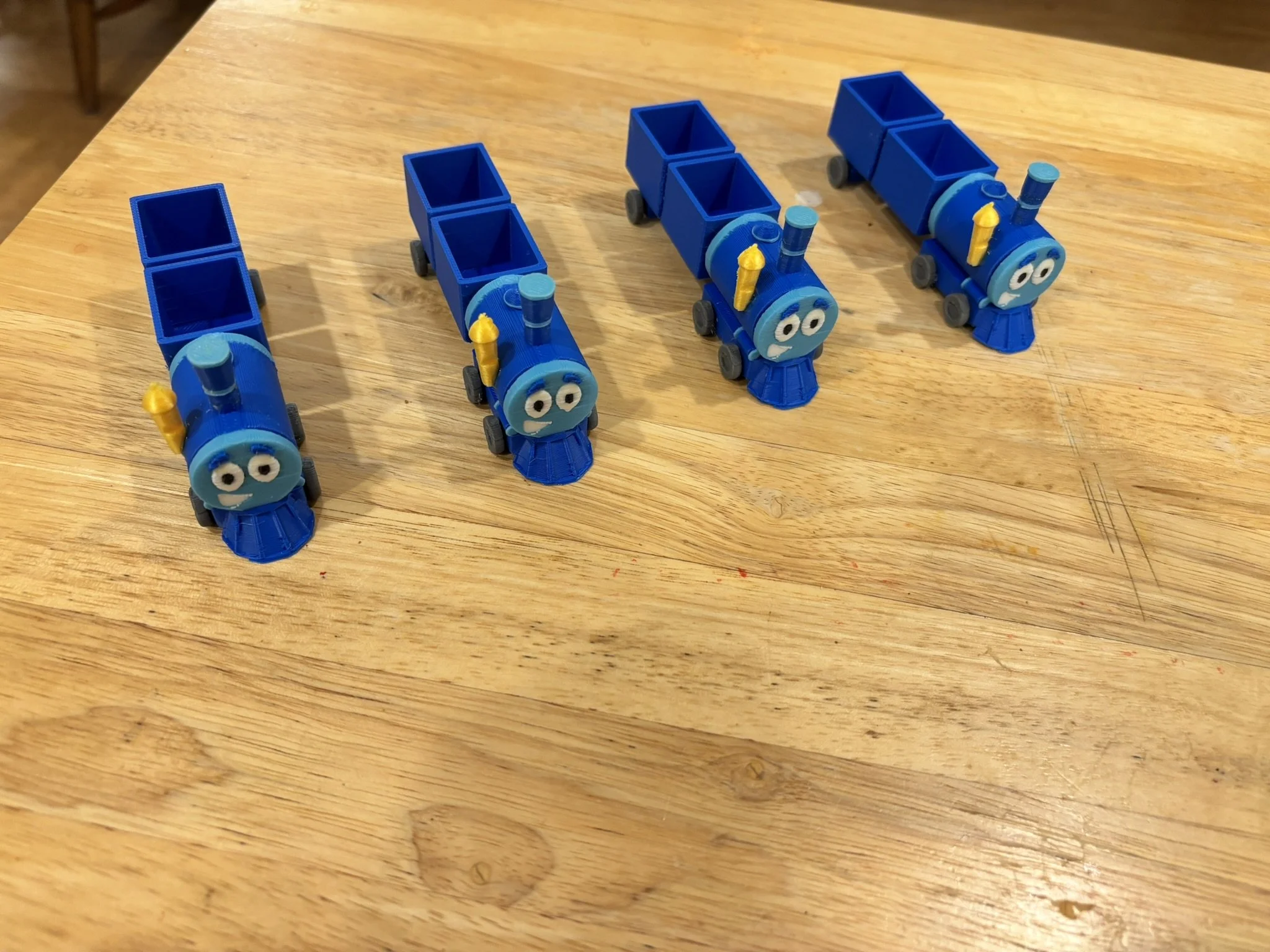Four small blue toy trains with cartoon faces and black wheels are lined up on a wooden surface.