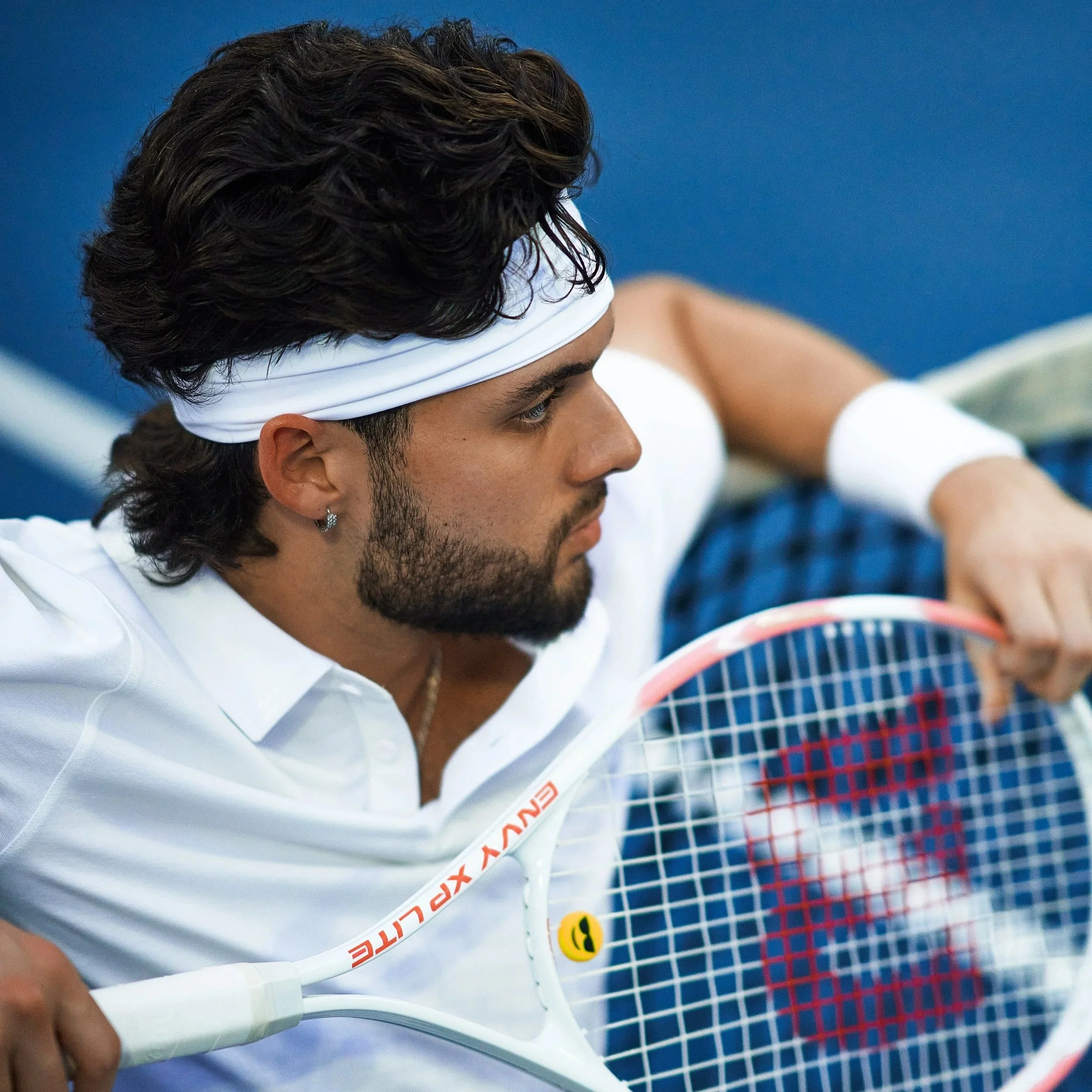 A male tennis player wearing a white headband and a white shirt, holding a tennis racket with a blue and red grid pattern, in a close-up shot. This amazing photo was captured and edited by Los Angeles-based Kairos VZN Premium Media Production.