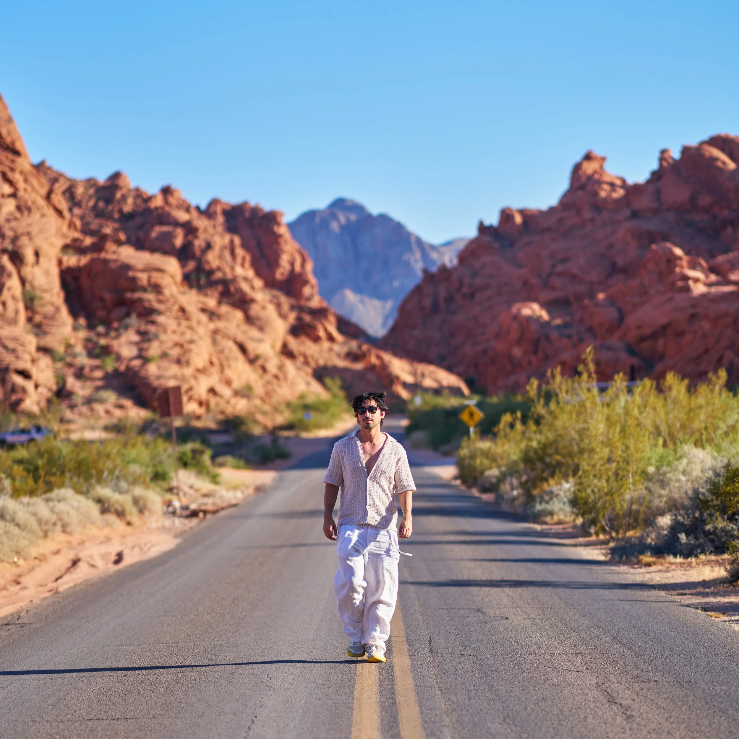 A young man in sunglasses walking on a deserted road in a desert landscape with red rock formations and mountains in the background on a clear day. This amazing photo was captured and edited by Los Angeles-based Kairos VZN Premium Media Production.