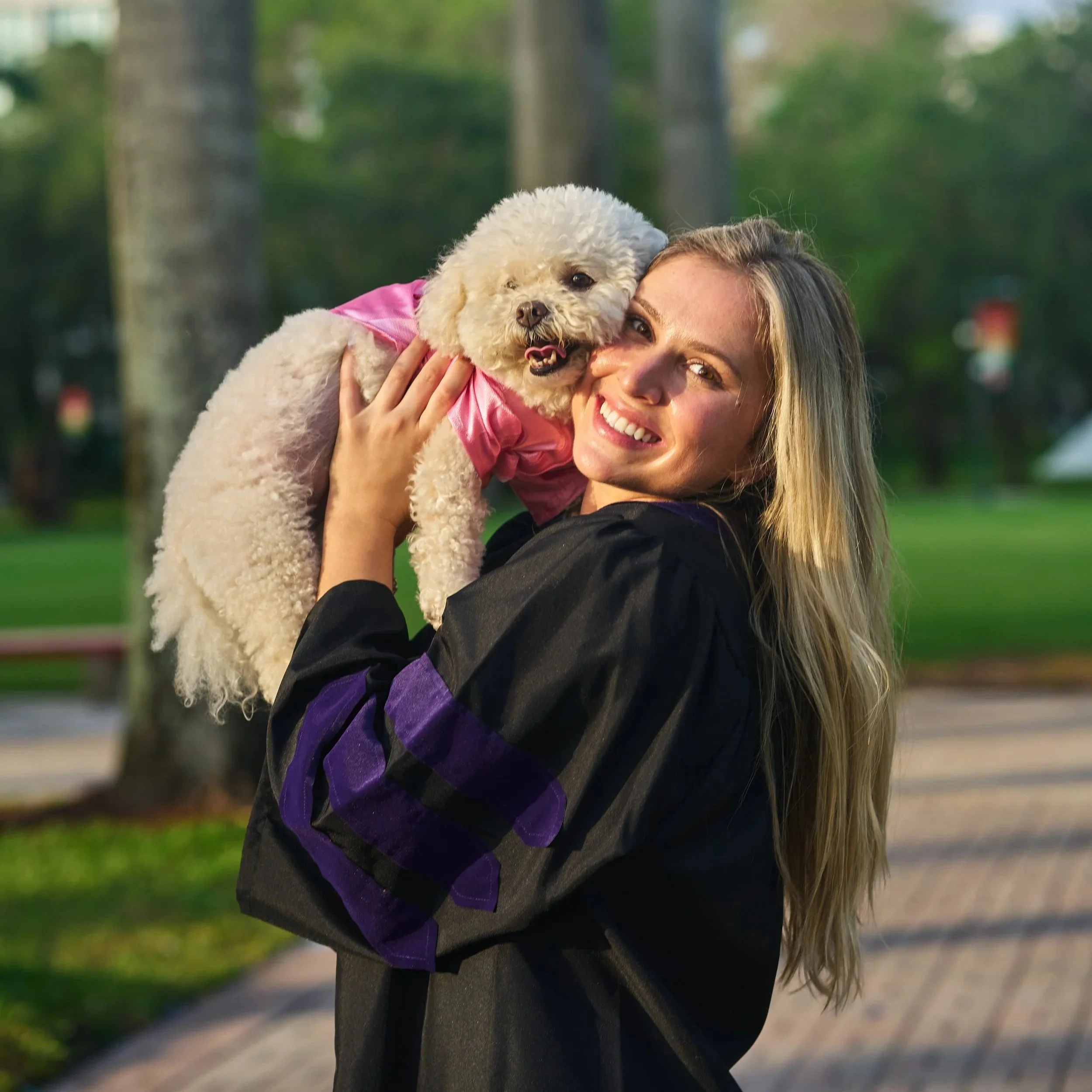 A college graduate in her graduation robe smiling and holding her dog, outdoors with trees in the background. This amazing graduation photo was captured and edited by Los Angeles-based Kairos VZN Premium Media Production.