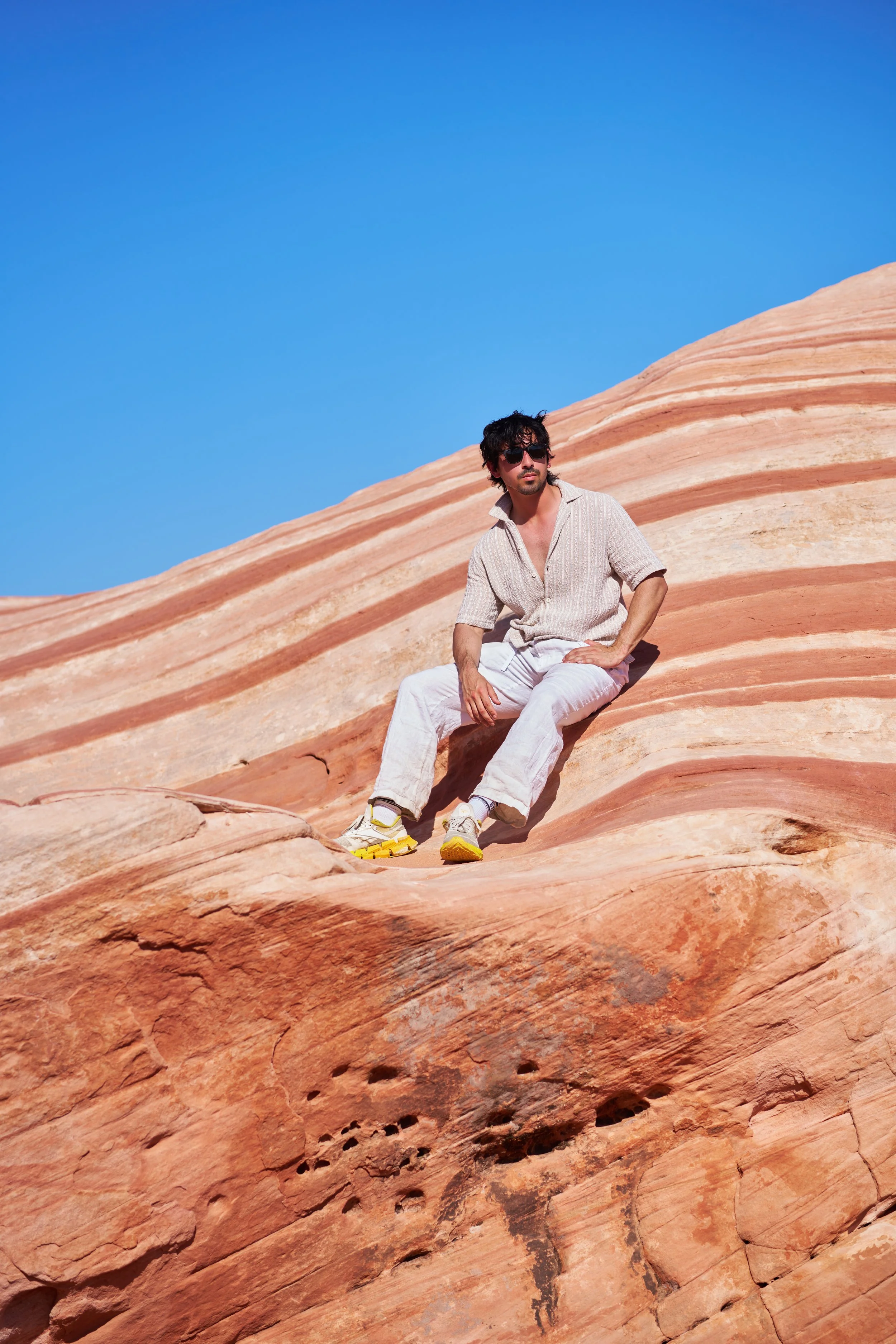 A picture of a man wearing sunglasses, a beige shirt, white pants, and yellow shoes sitting on orange sandstone formations beneath a clear blue sky. The amazing photo was captured and edited by Los Angeles-based Kairos VZN Premium Media Production.