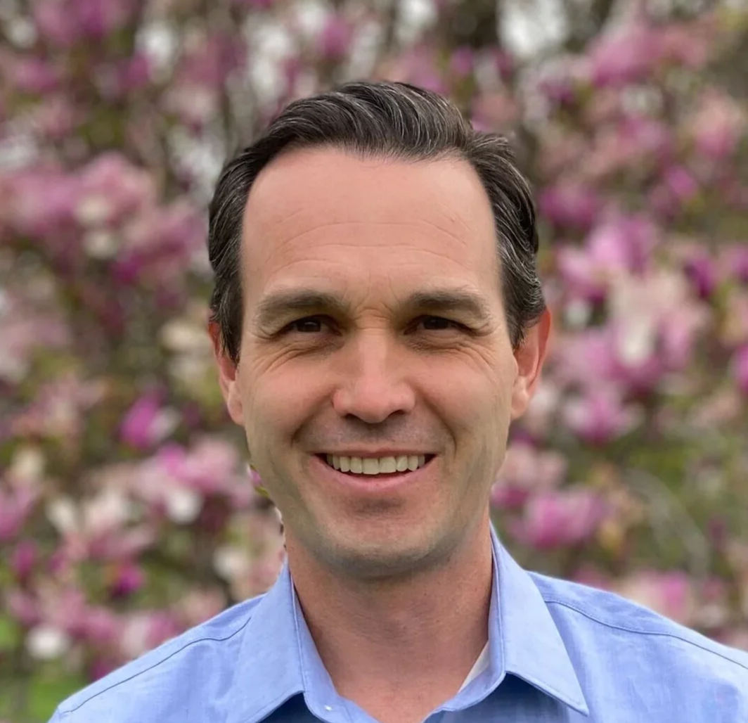 A smiling man with dark brown hair in a blue collared shirt stands outdoors in front of a blurred background of pink flowering trees.