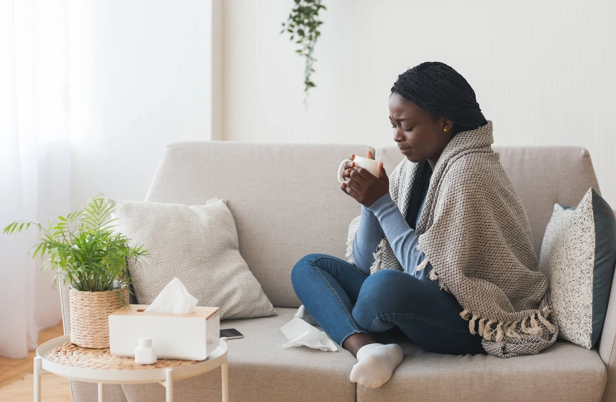 A woman sitting on a beige couch, holding a mug with both hands, with tissue papers around her, looking distressed or unwell, in a bright living room with plants and cushions.